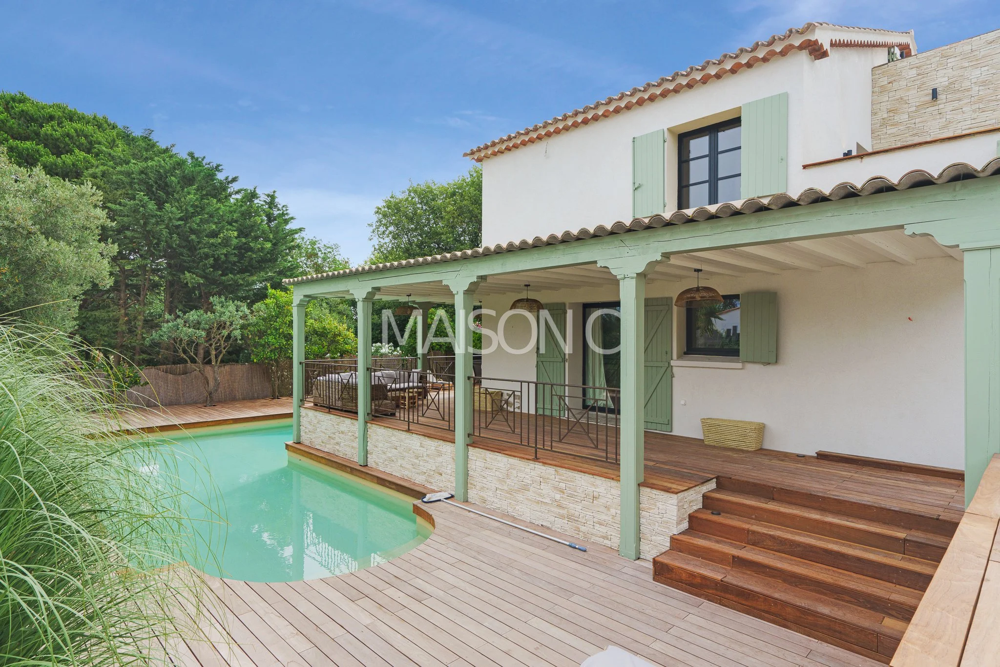 Maison avec terrasse en bois, piscine, et véranda, entourée de verdure, sous un ciel bleu.