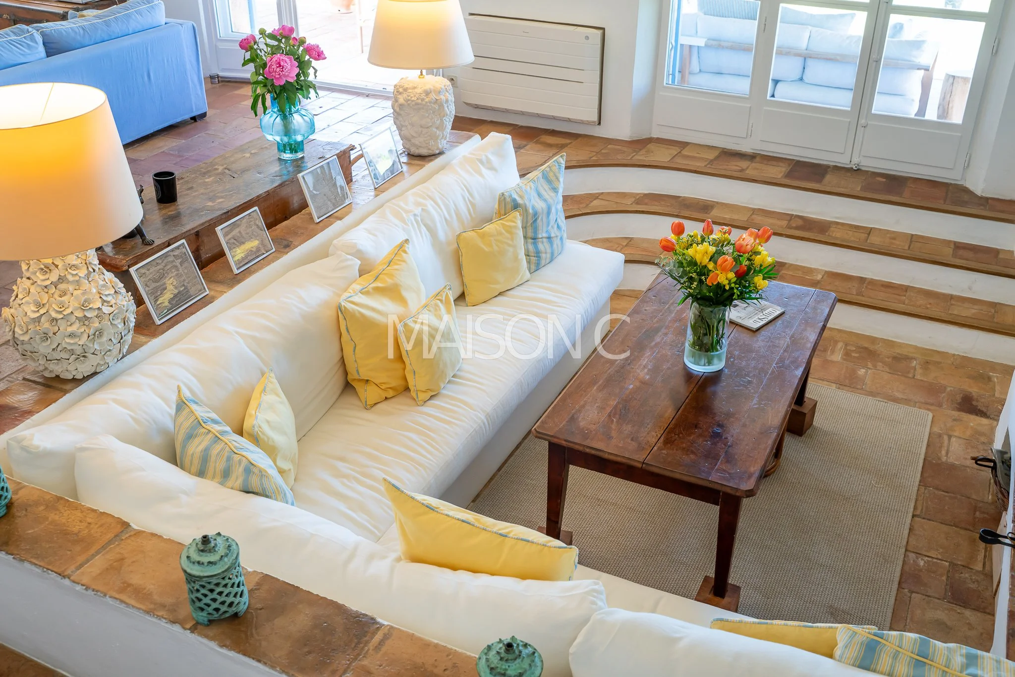 Salon avec canapé blanc, coussins jaunes et bleus, table en bois avec un vase de fleurs colorées, lampes, fenêtres, et décorations