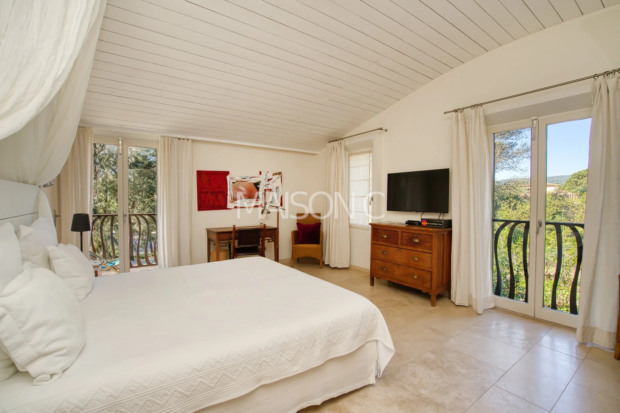 Chambre avec lit blanc, télévision, fenêtres avec vue sur nature, mobilier en bois, décoration simple, rideaux blancs, sol en pierre