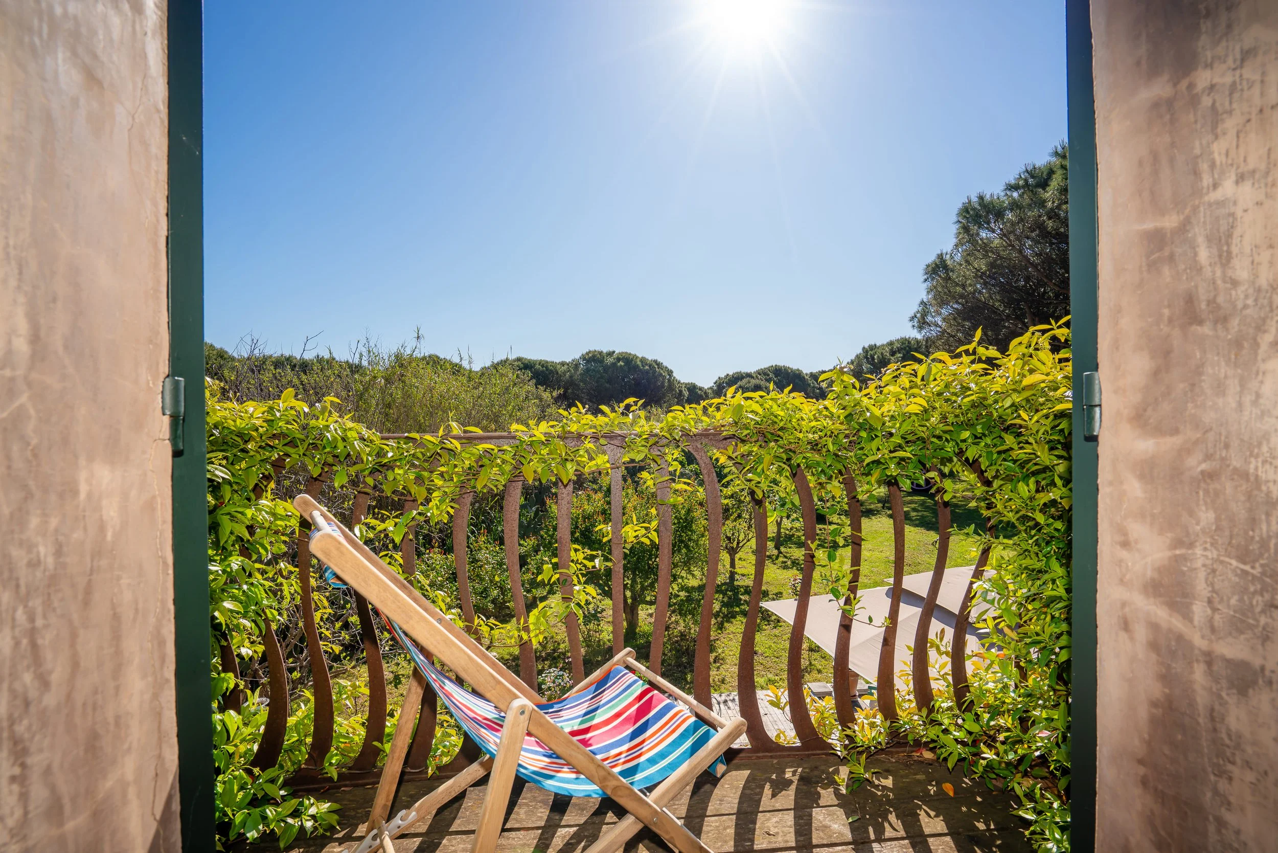 Balcon avec un fauteuil pliante en bois et toile à rayures colorées, entouré de végétation, vue sur un jardin et un ciel ensoleillé.
