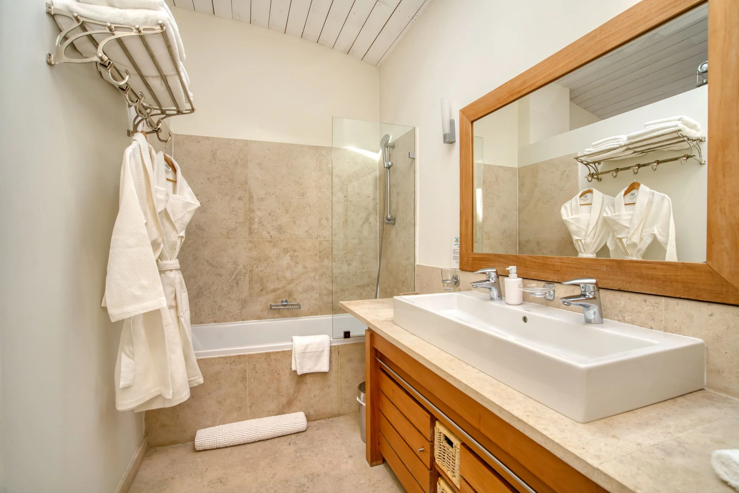 Salle de bain avec un lavabo, un grand miroir en cadre en bois, une baignoire avec une paroi en verre, un porte-serviettes avec serviettes, et des robes blanches suspendues.