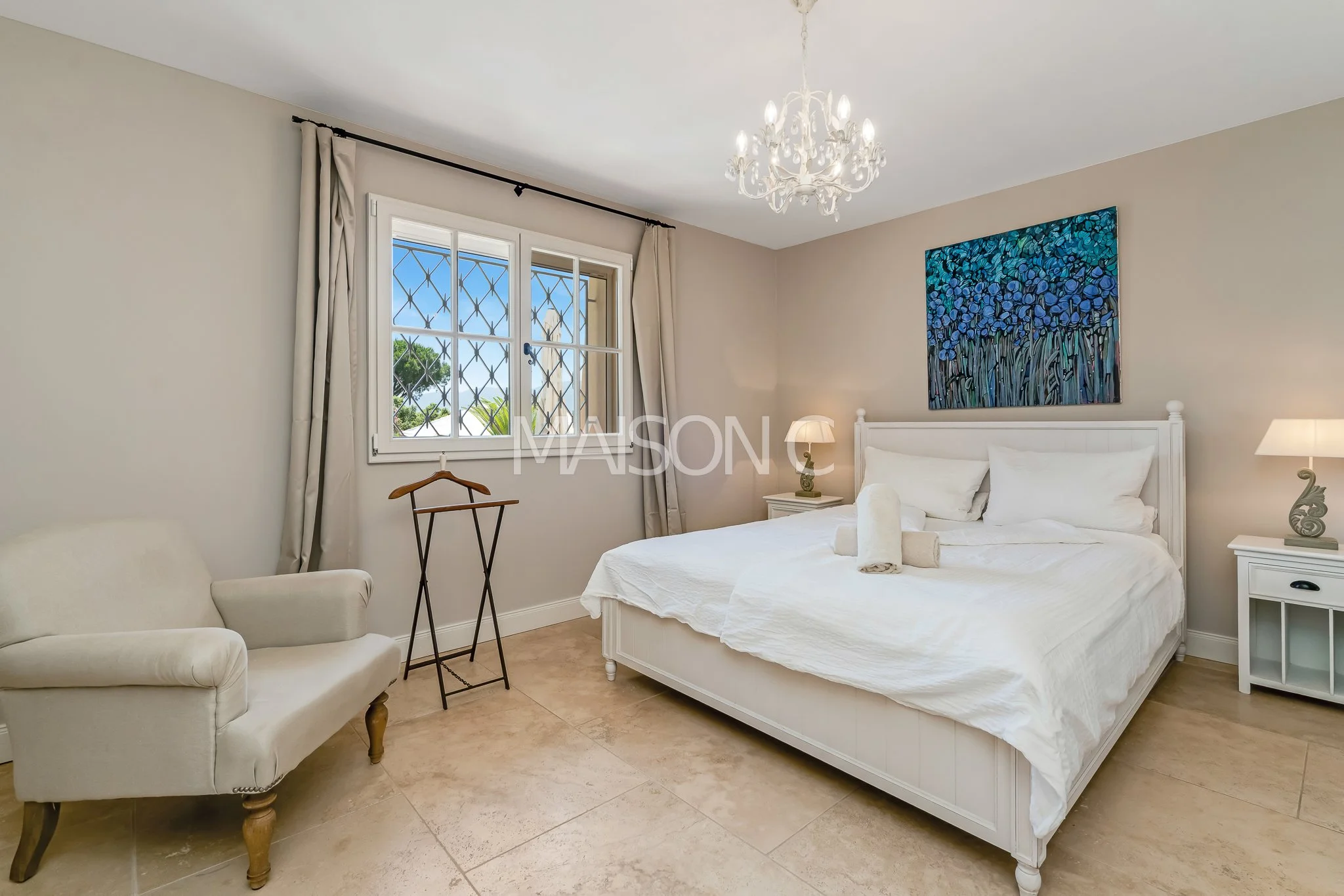 Chambre lumineuse avec lit blanc, tableau de fleurs bleues, deux lampes de chevet, fauteuil beige, fenêtre avec barre de sécurité et rideaux blancs, lustre blanc.