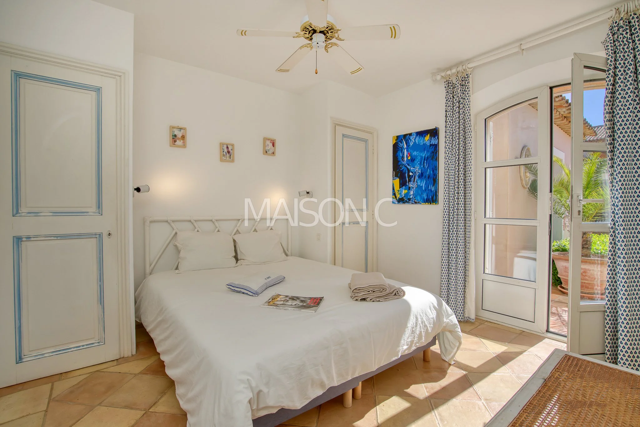 Chambre lumineuse avec lit blanc, porte-fenêtre donnant sur un balcon avec plantes, décorée de tableaux modernes et de rideaux à motifs bleus.