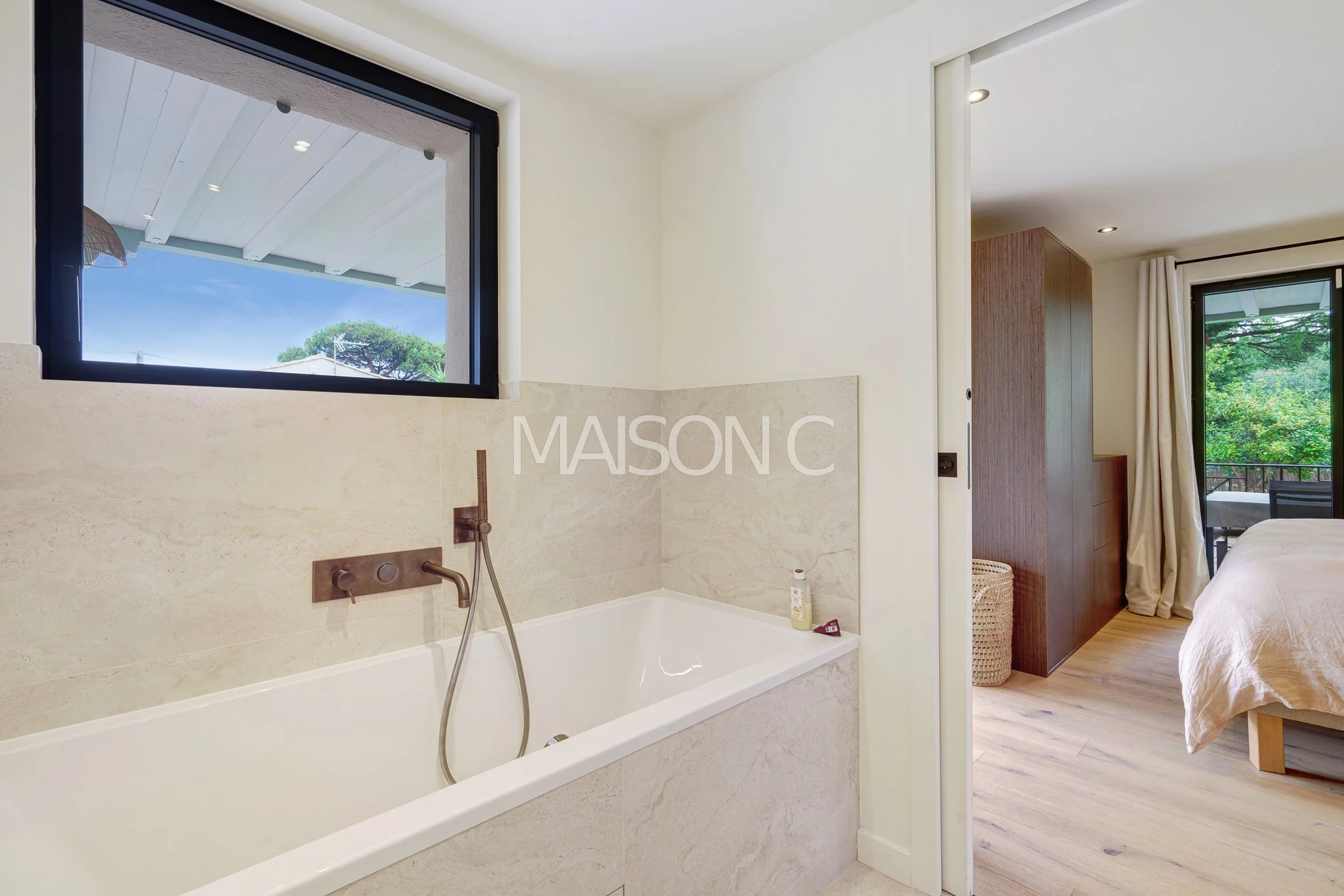 Salle de bain attenante à une chambre, vue intérieure, baignoire en marbre, grande fenêtre avec vue sur le ciel et la nature, porte de la chambre entrouverte, parquet en bois, décoration moderne et lumineuse