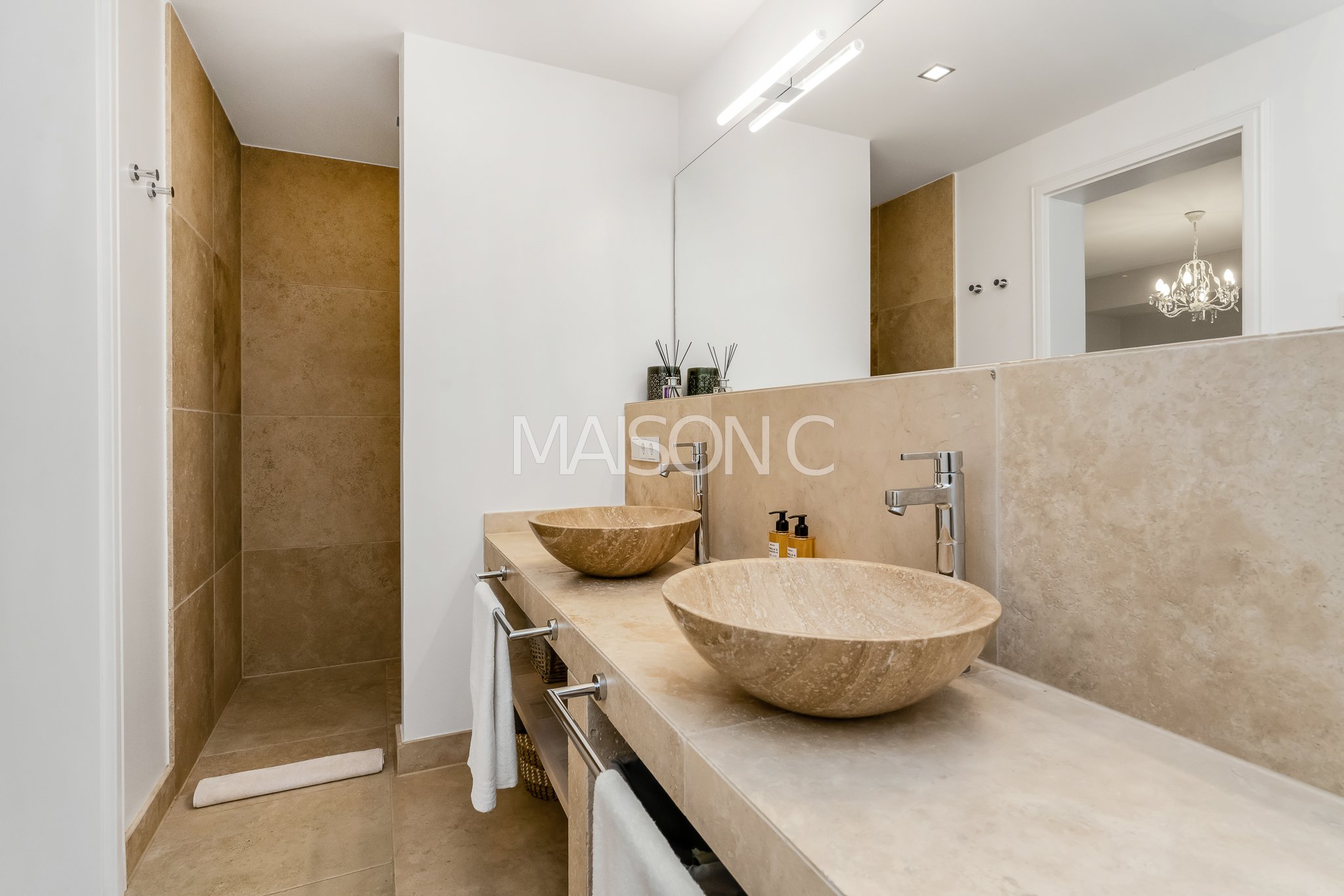 Salle de bain avec double lavabo en pierre beige, miroir mural, éclairage moderne, et espace de douche en arrière-plan.