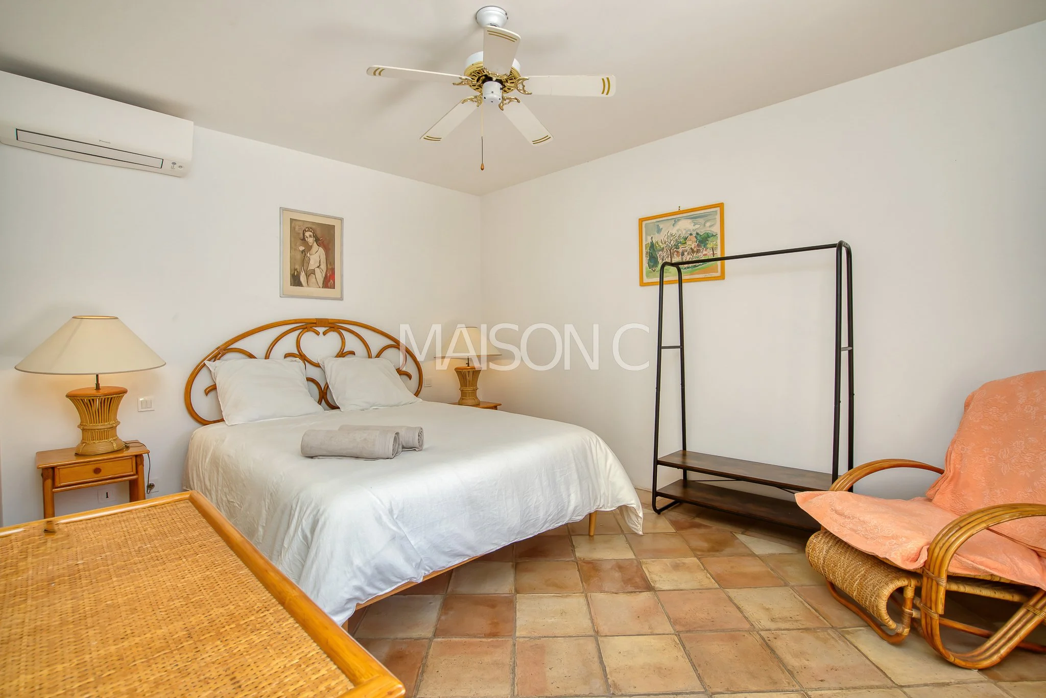 Chambre avec un lit, une table de chevet, une lampe, un tableau, un fauteuil en rotin avec un coussin, un arbre à vêtement, un plafond avec un ventilateur et un climatiseur, sol en carreaux de terre cuite.