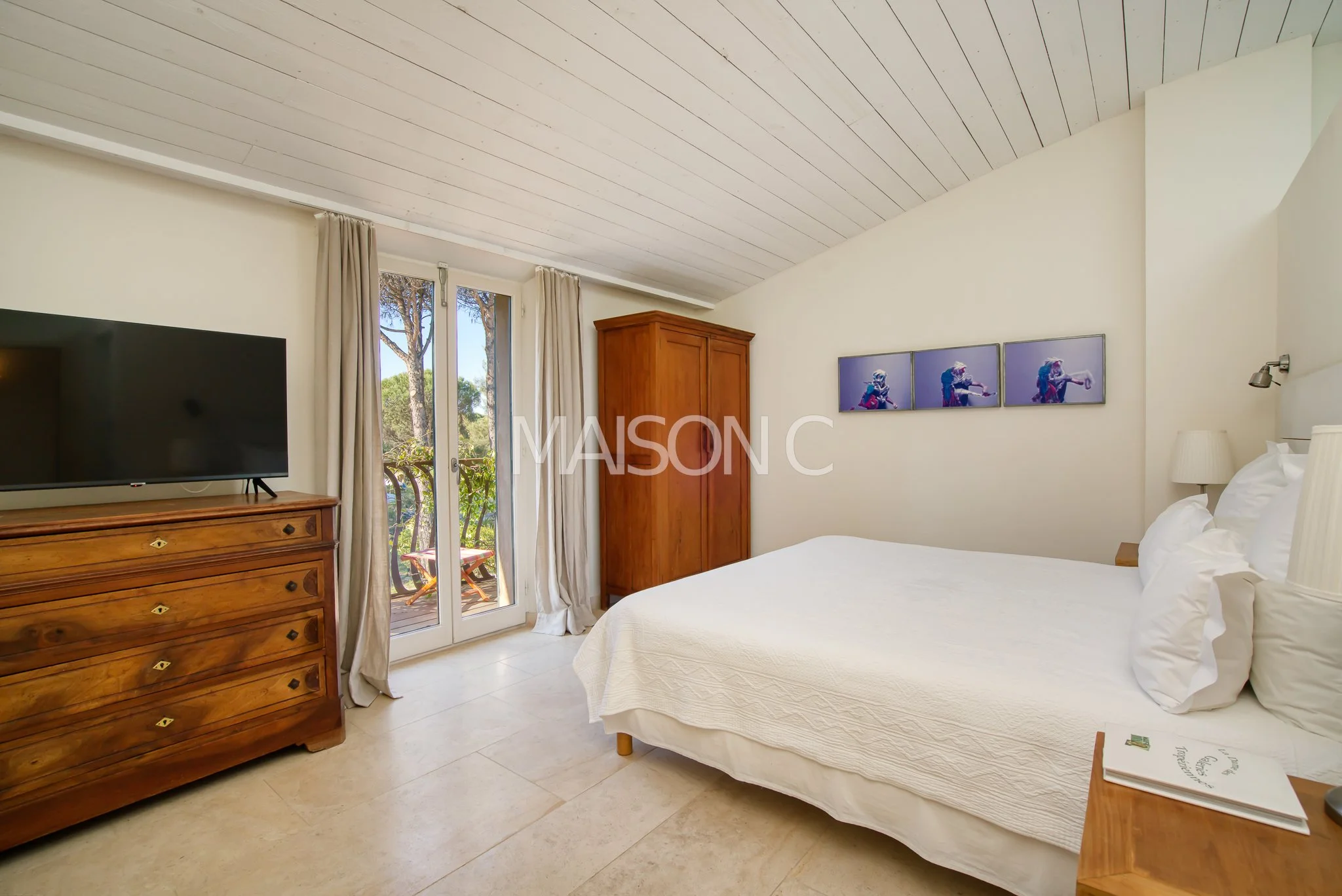 Chambre lumineuse avec un lit blanc, une armoire en bois, une commode avec une télévision, une porte-fenêtre donnant sur un balcon avec des volets en bois, et une décoration moderne sur le mur