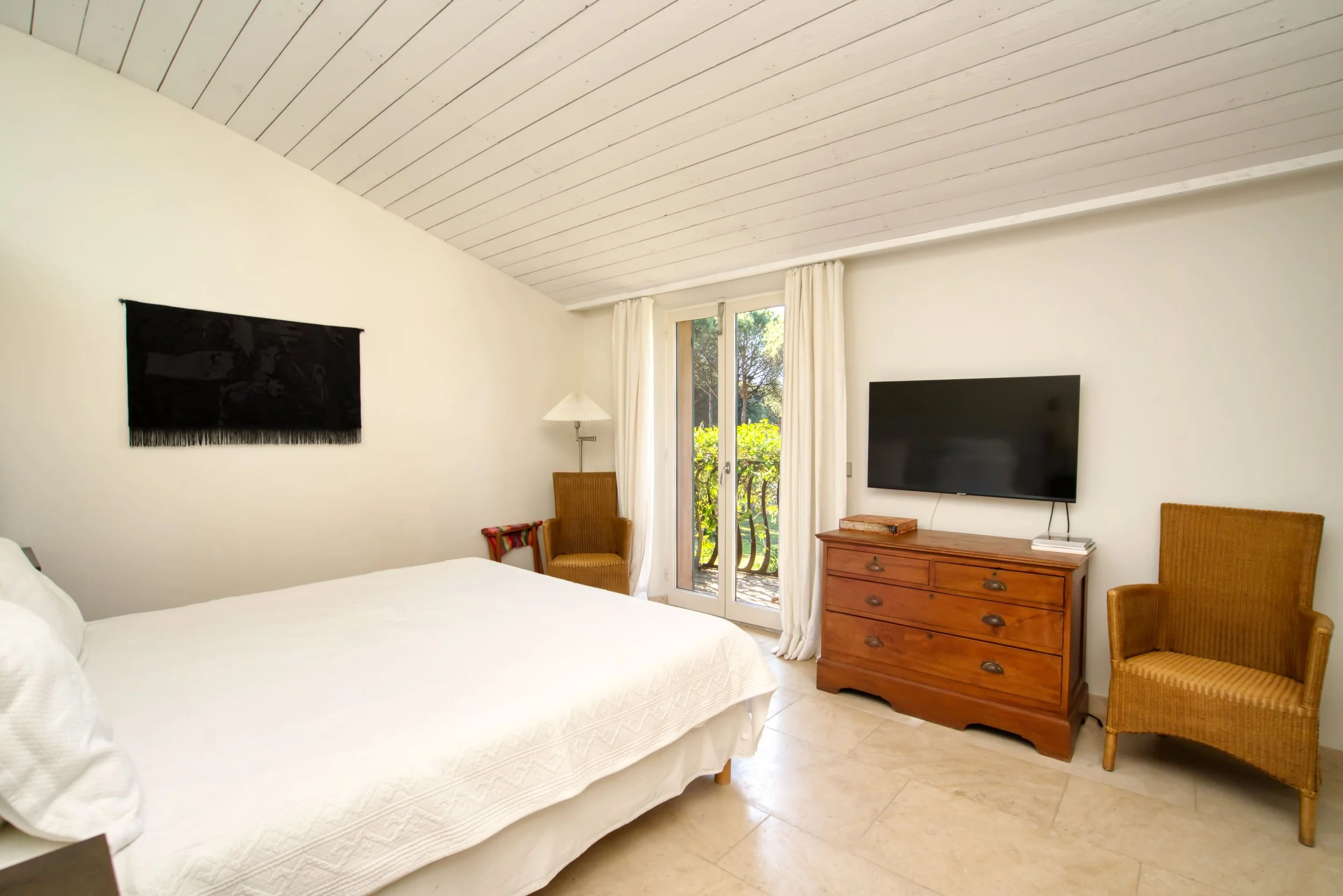 Chambre de lit avec mur blanc, télévision à écran plat, meubles en bois, porte-fenêtre donnant sur un balcon avec vue sur la nature, fauteuils en rotin, lampe de lecture, décoration simple.