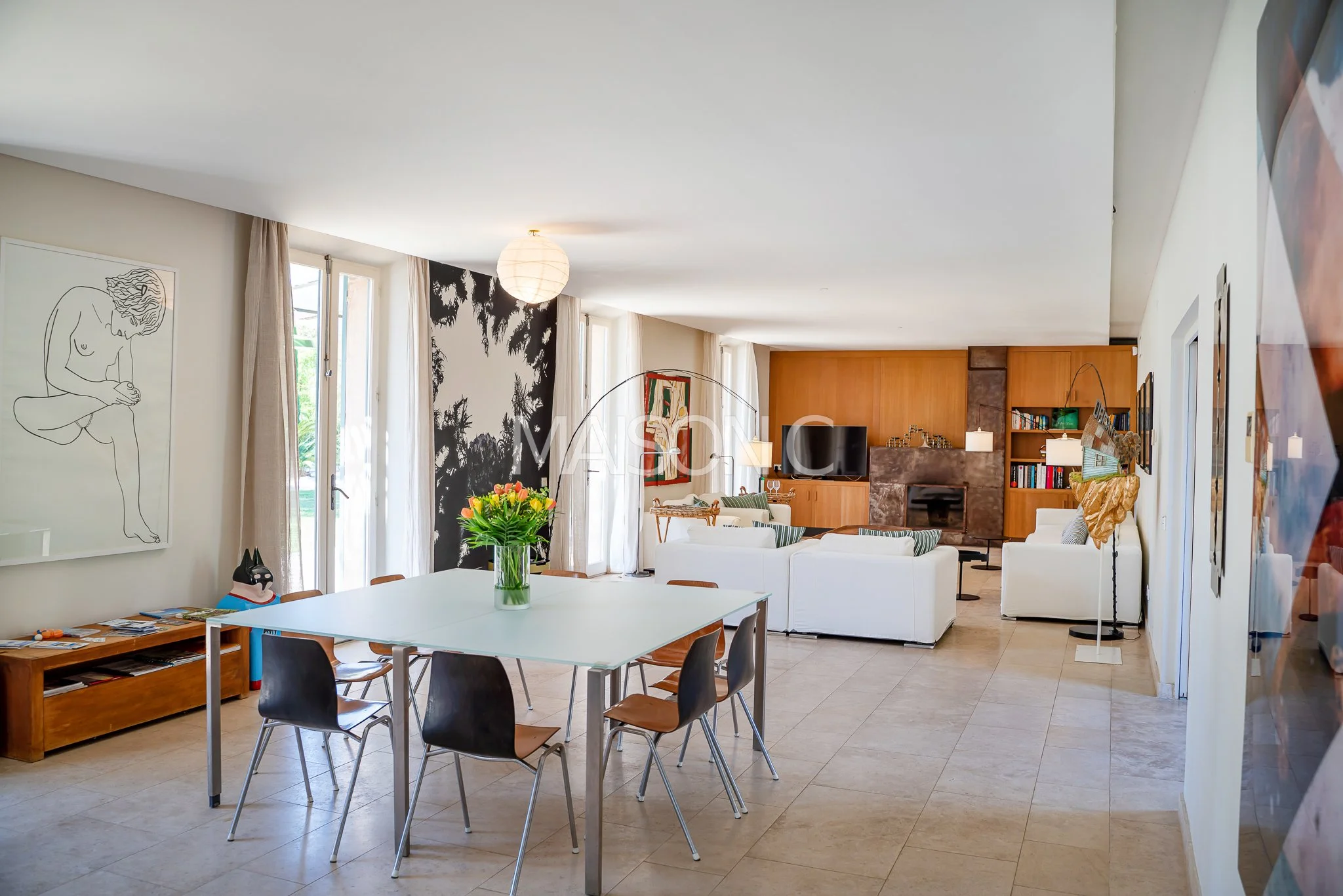 Salon spacieux avec un canapé blanc, une cheminée en pierre, une bibliothèque, un tapis, une télévision, et une décoration moderne. Espace de vie lumineux avec grandes fenêtres et rideaux clairs.