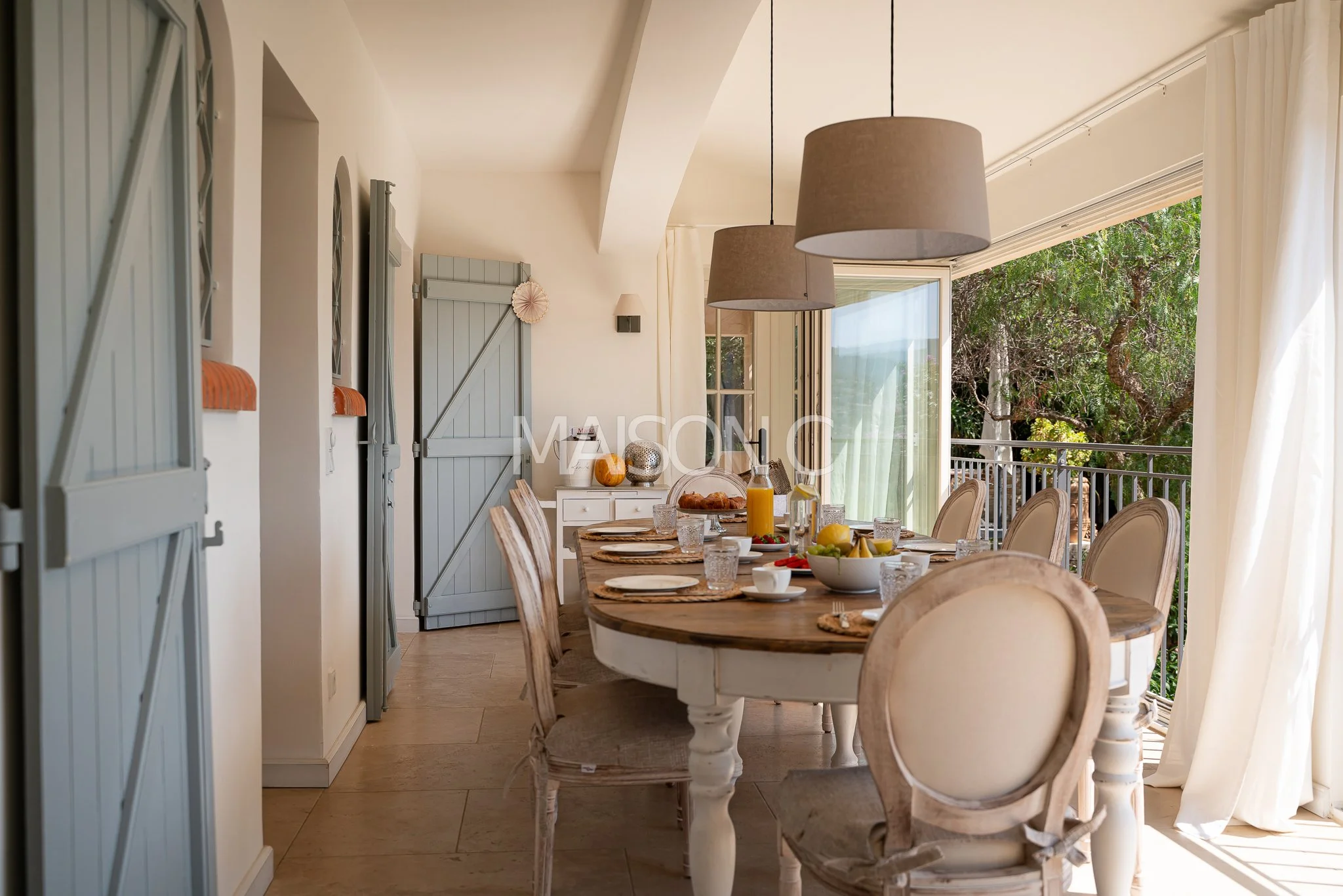 Salle à manger avec une table ronde en bois, chaises beige, fruits, boissons, grande baie vitrée ouverte sur un balcon avec vue sur des arbres verdoyants, éclairage suspendu