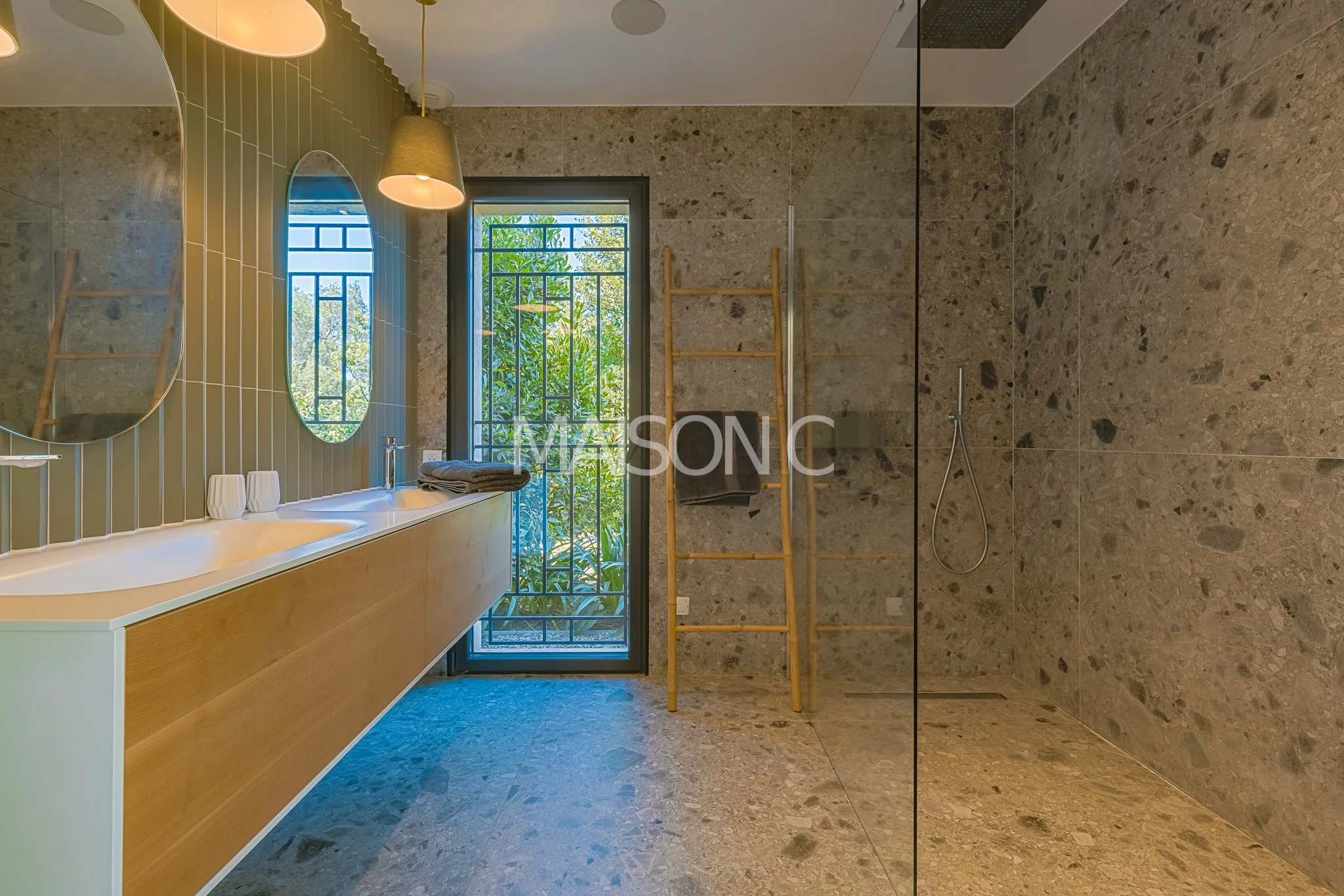 Salle de bain moderne avec double lavabo, deux miroirs ovales, grande fenêtre avec vue sur la nature, porte en verre menant à l'extérieur, porte-manteaux en bois, serviettes encadrées, sol et murs en pierre.