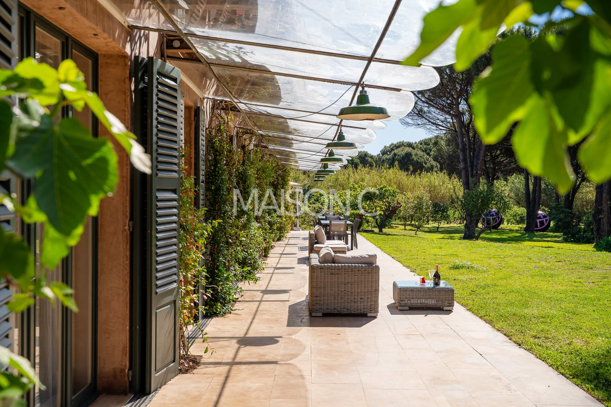 Terrasse extérieure avec mobilier en rotin, vue sur un jardin arboré ensoleillé, sous un auvent transparent avec des lampes suspendues, entourée de végétation luxuriante.