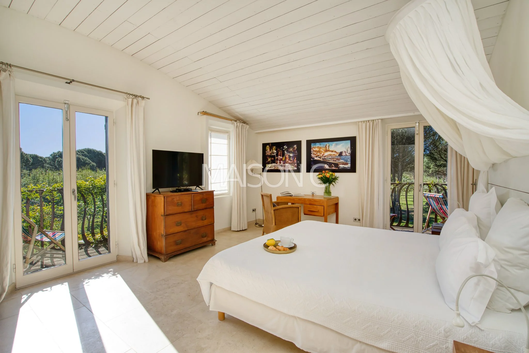 Chambre lumineuse avec un lit blanc, une télévision sur un meuble en bois, un bureau avec une chaise, deux portes fenêtres avec des rideaux blancs, vue sur un jardin, décorée avec des photos encadrées et un bouquet de fleurs.