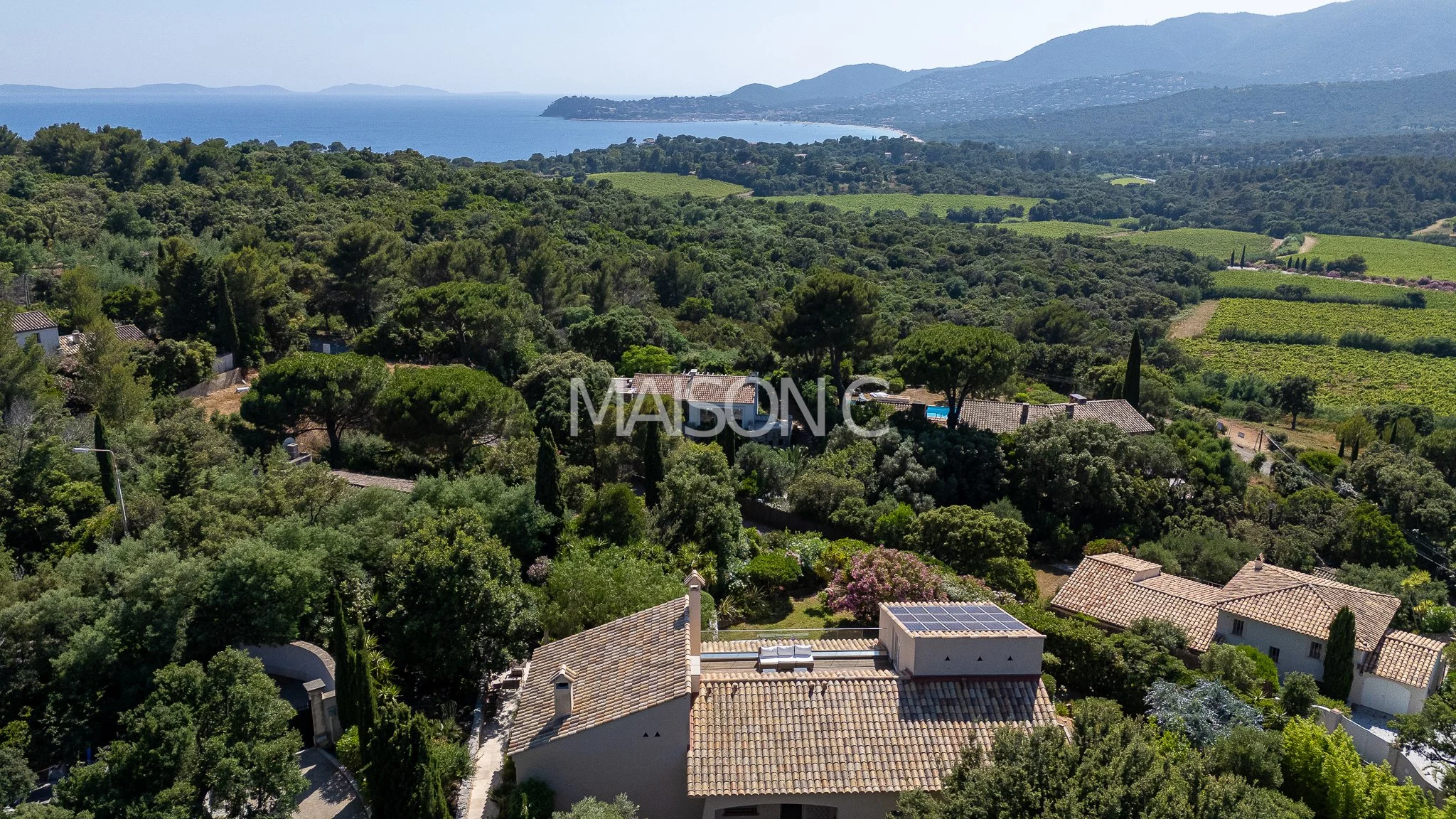 Vue aérienne de maisons avec toits en tuiles dans une zone verdoyante, avec la mer et des montagnes en arrière-plan.