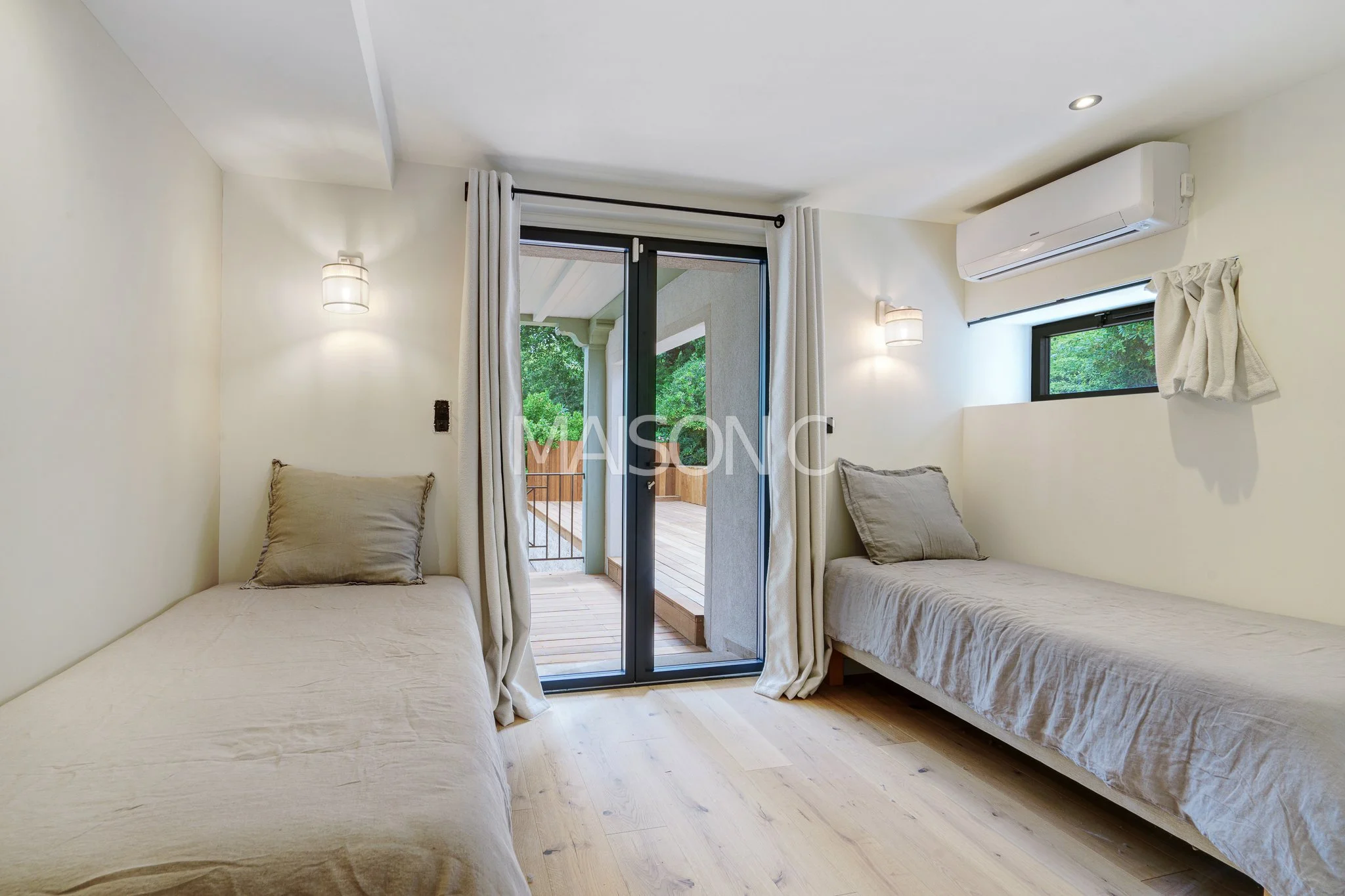 Chambre avec deux lits simples, grande fenêtre, porte-fenêtre donnant sur un balcon en bois, murs blancs, rideaux blancs, climatiseur, éclairage encastré