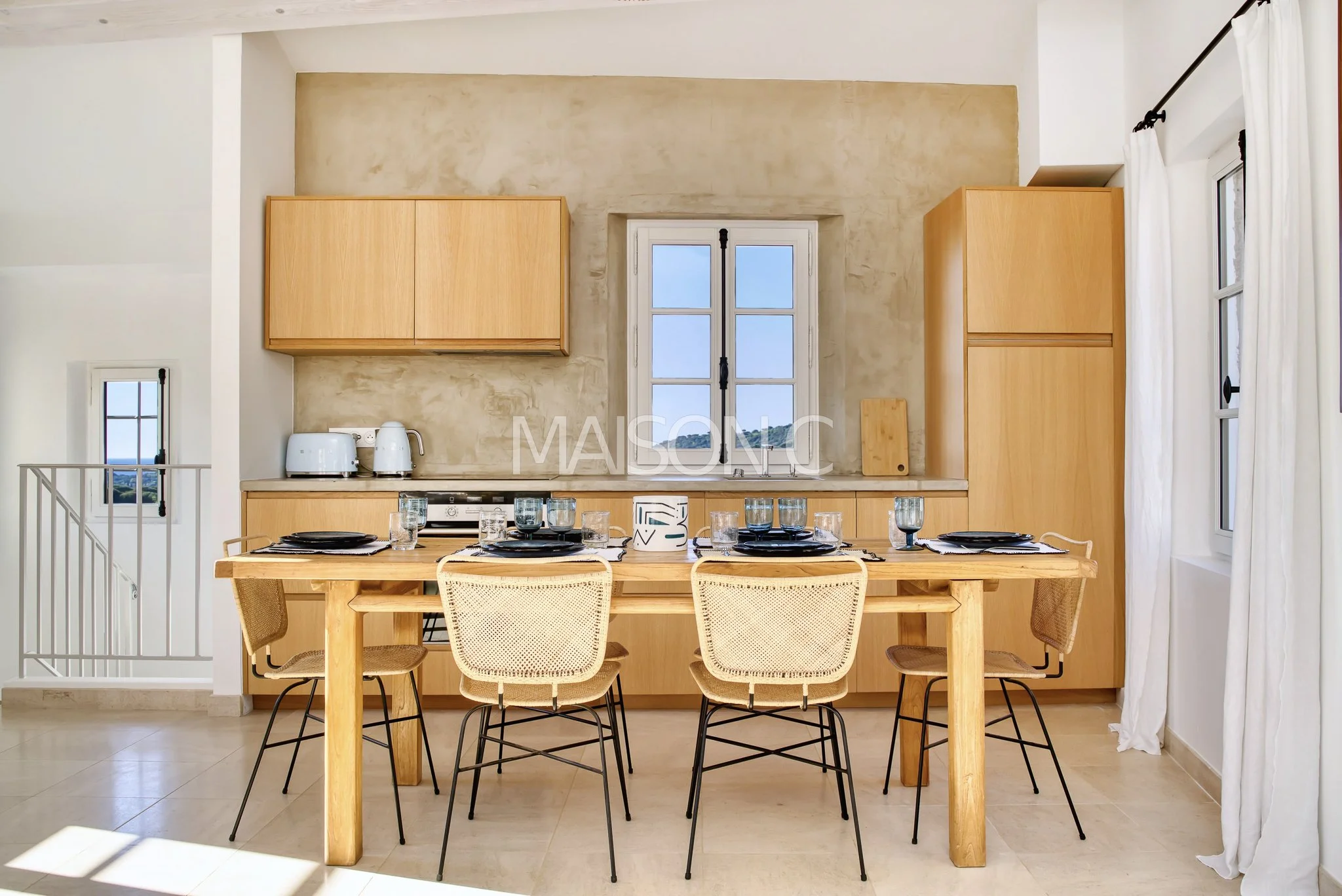 Salle à manger avec table en bois, six chaises, cuisine en arrière-plan, deux petits placards en bois, fenêtre avec rideaux blancs, sol en carrelage beige, mur en plâtre clair.