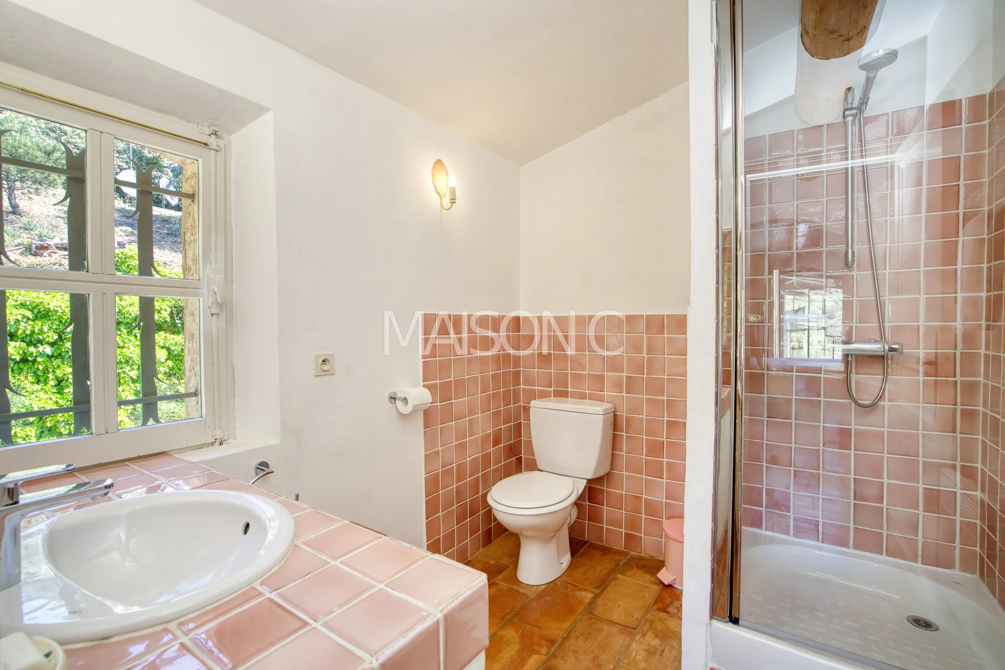 Salle de bain avec un lavabo, un WC, et une cabine de douche, murs et sol en carreaux roses, fenêtre laissant entrer la lumière naturelle.