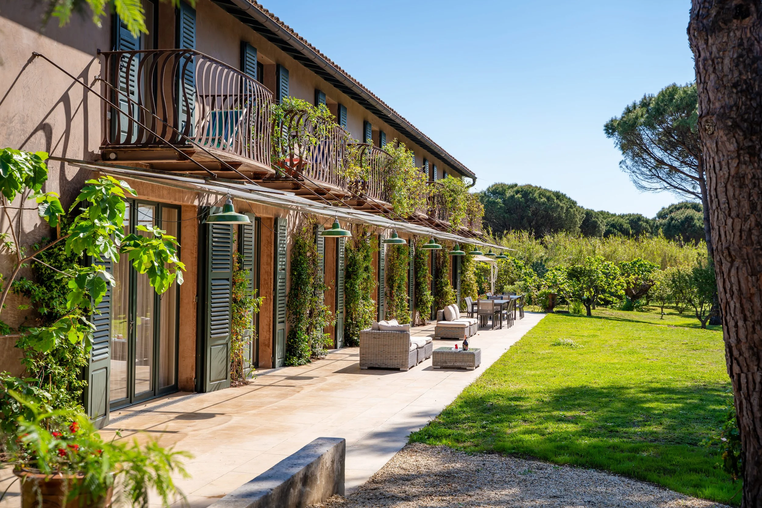 Facade d'une maison avec terrasse et salon extérieur, entourée de végétation et d'arbres, sous un ciel bleu clair.