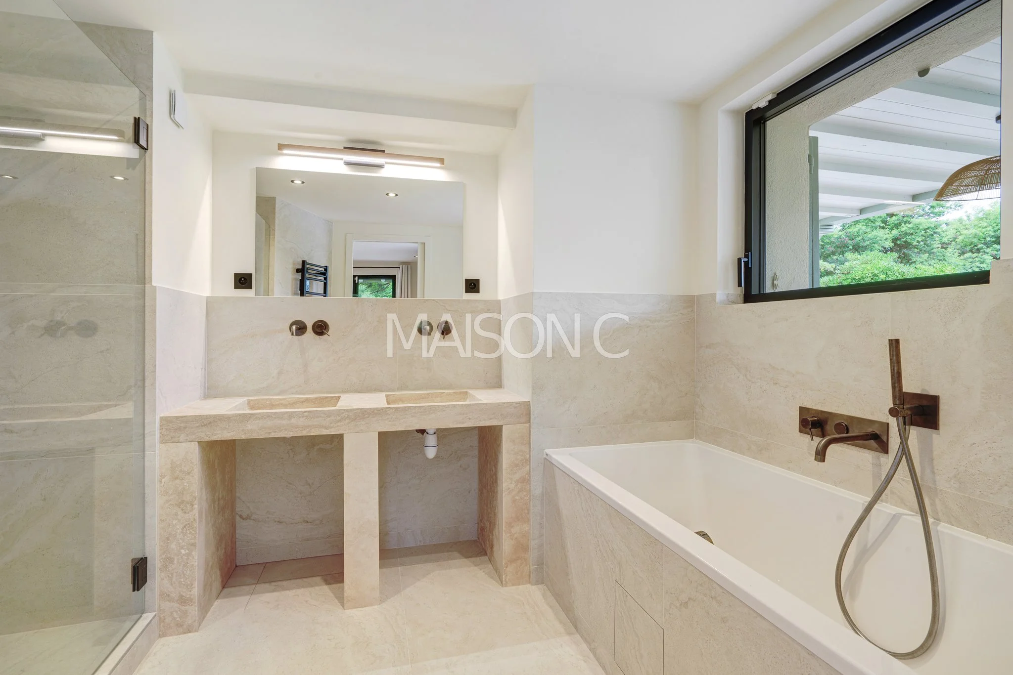 Salle de bain moderne avec une baignoire, une double vasque en pierre, un miroir, une fenêtre ouvrante avec vue sur la nature, et un éclairage intégré.