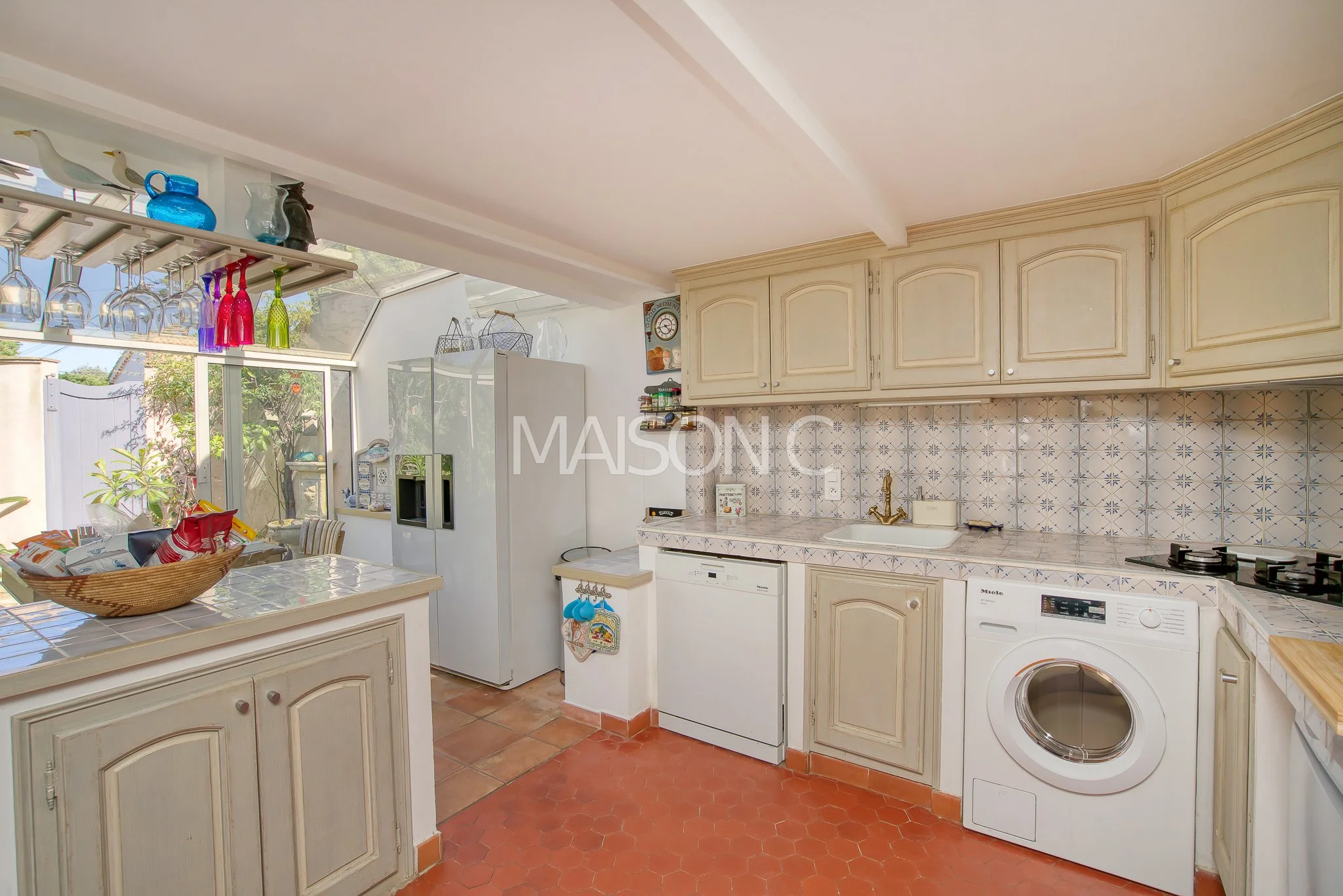 Cuisine lumineuse avec appareils électroménagers intégrés, armoire en bois clair, mur de carreaux bleu et blanc, accès à un espace extérieur avec lacets et plantes