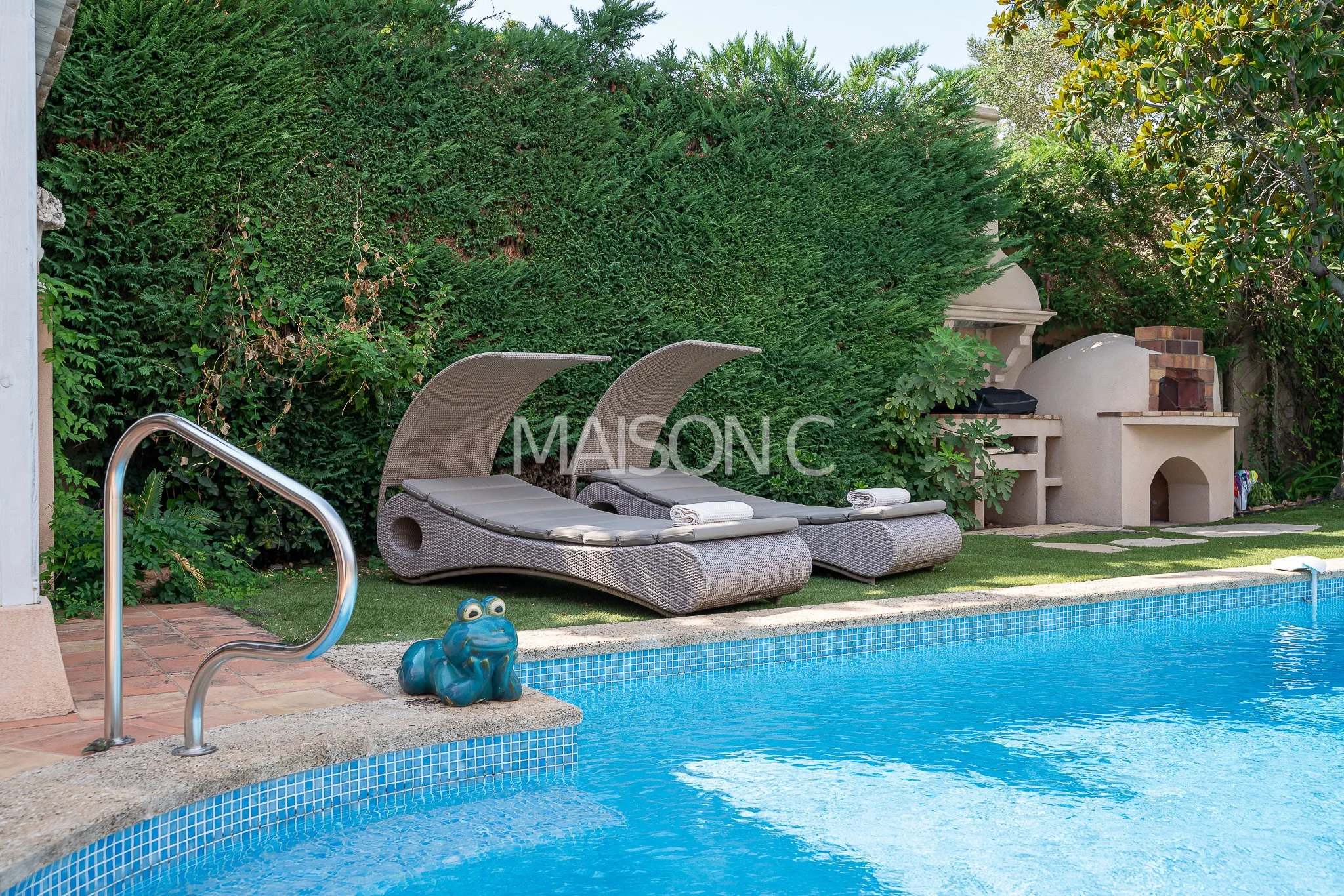 Deux fauteuils de soleil en rotin avec des coussins gris, une petite sculpture de grenouille bleue en plastique, une piscine bleue avec un escalier en métal, un barbecue en pierre blanc, un mur de haies vertes et une pelouse.