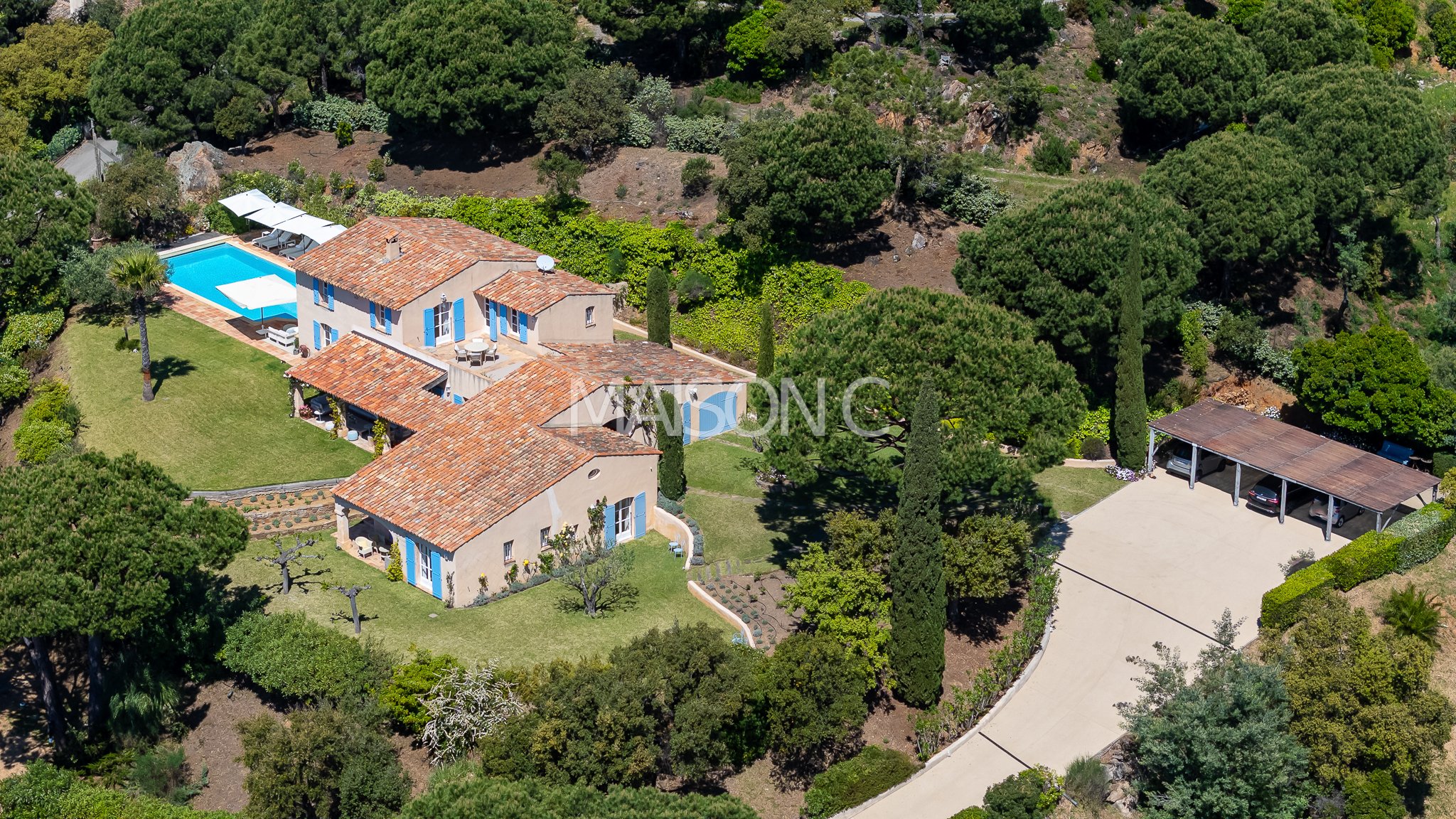 Vue aérienne d'une grande maison de style méditerranéen avec cheminée en terre cuite, piscine, terrasse aménagée et jardin entouré d'arbres. Un garage avec véhicules est visible à droite.