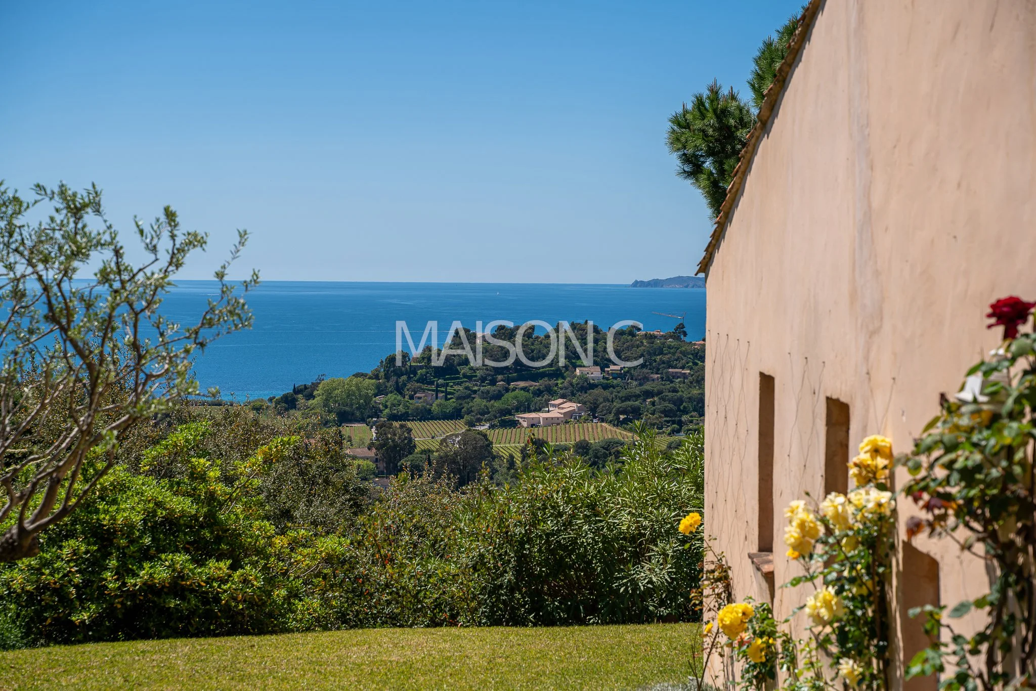 Vue d'une maison en face de la mer avec des plantes et des fleurs colorées, un ciel clair et une côte lointaine.