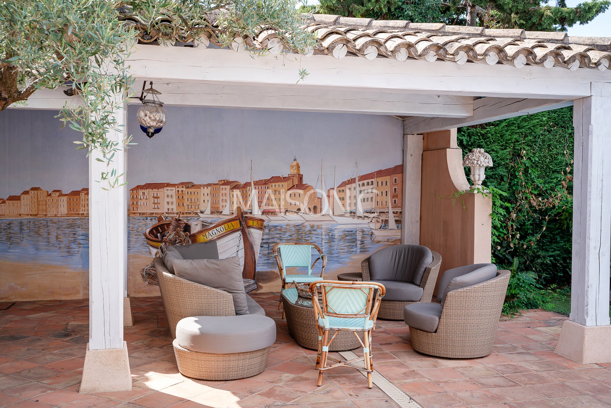 Une terrasse couverte avec plusieurs fauteuils et chaises en rotin et coussins, un mur peint avec une scène de port de plaisance, un lampadaire suspendu, et des plantes vertes en arrière-plan.