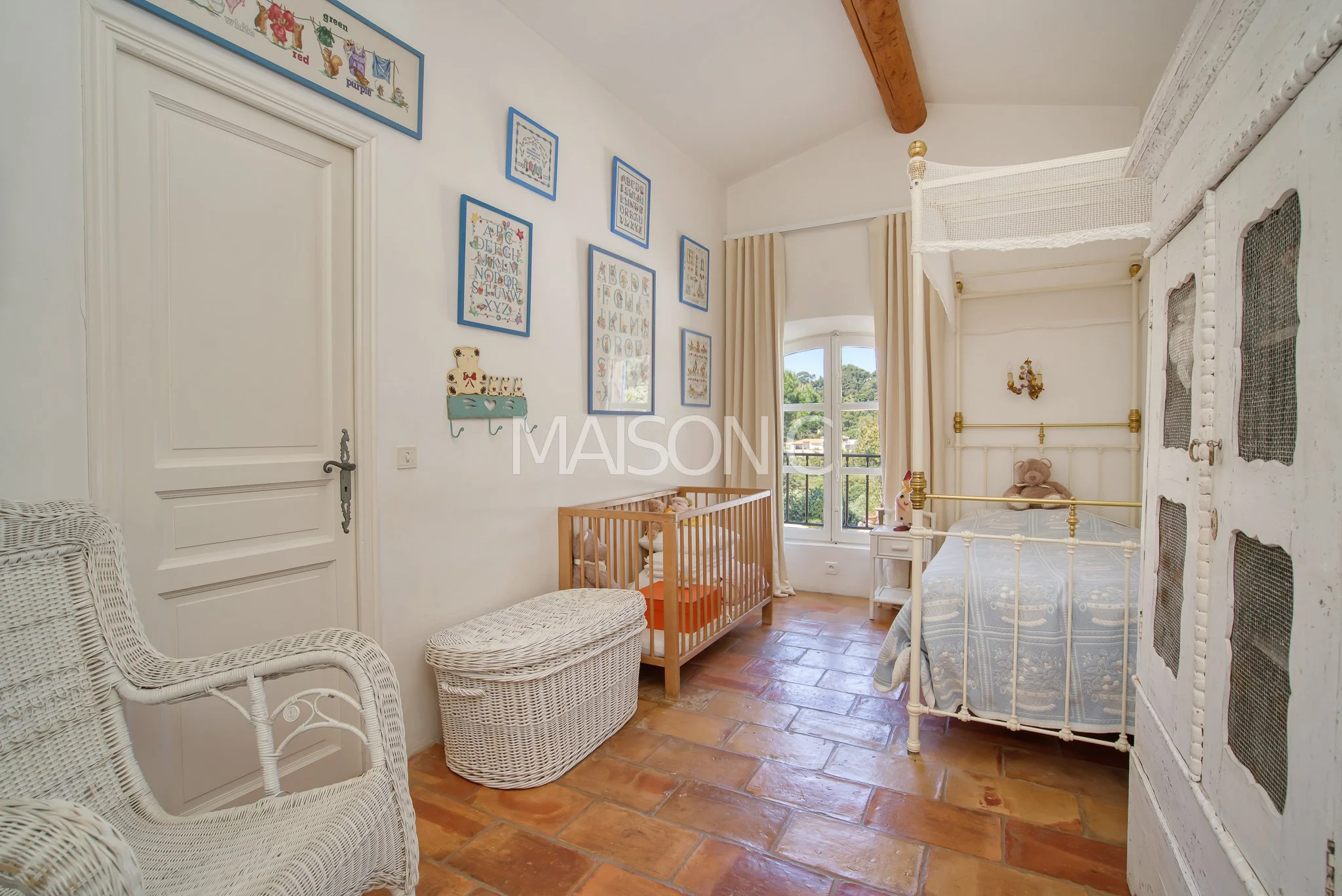 Chambre d'enfant avec lit à baldaquin, berceau, fauteuil en rotin, coffre de rangement en rotin, bibliothèque murale avec cadre coloré, grande fenêtre avec rideaux, décorations murales, sol en carrelage, ambiance lumineuse et douce.