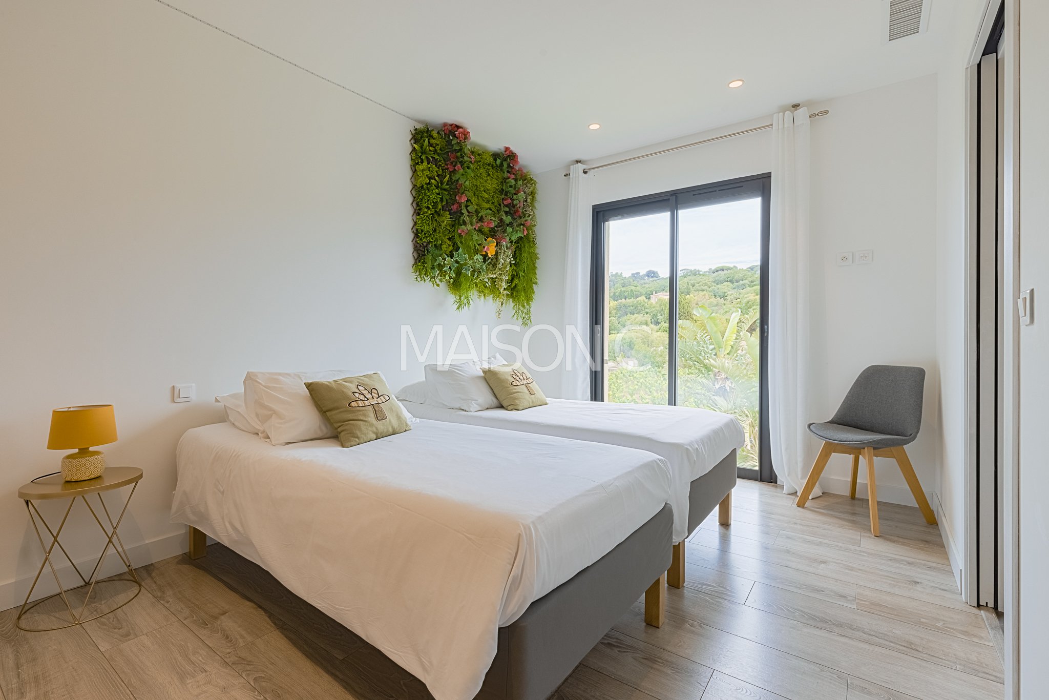 Chambre moderne avec deux lits, mur végétal décoratif, grande fenêtre donnant sur la nature, fauteuil gris, petite table de chevet avec lampe