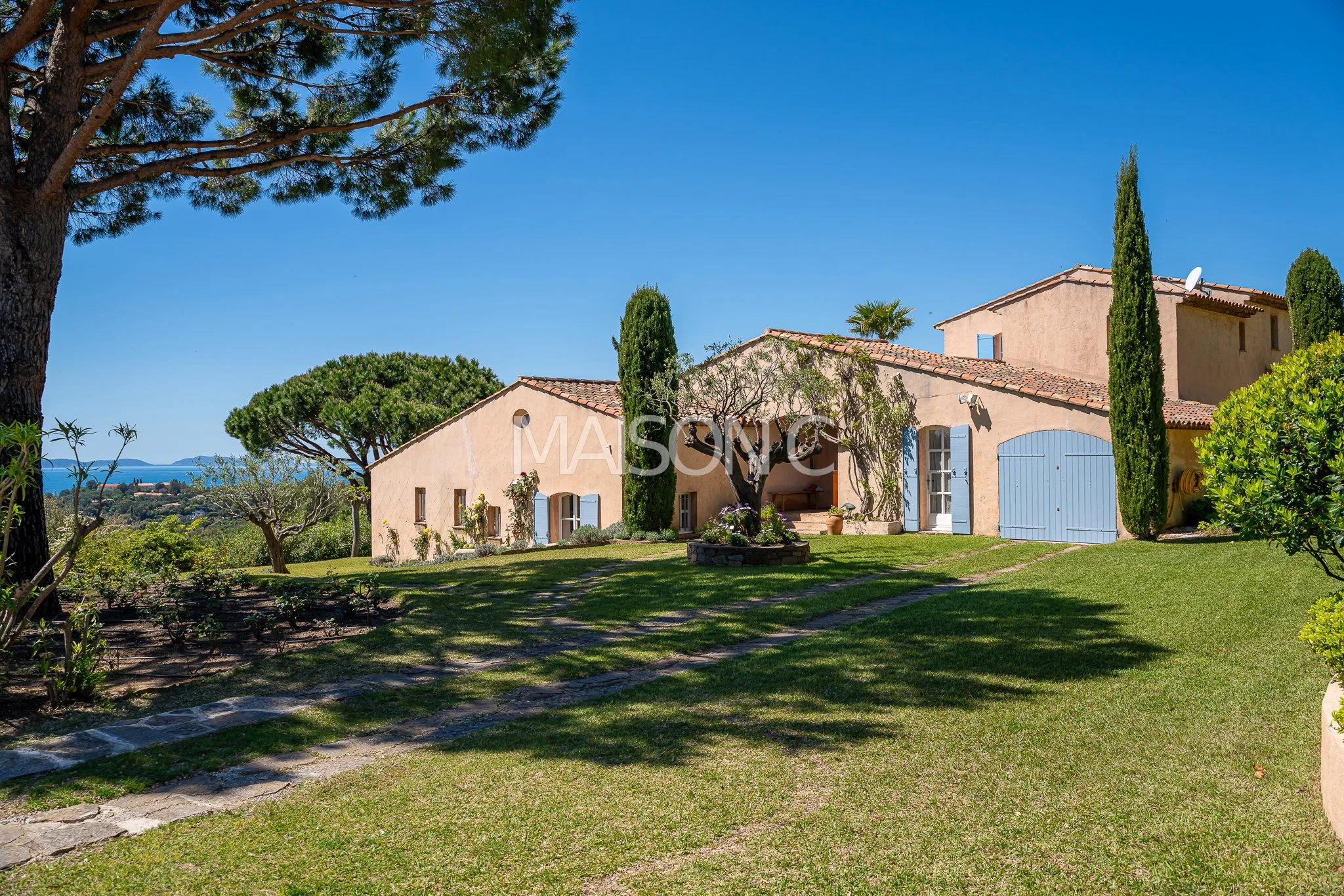 Maison de style provençal avec murs en stuc beige, volets bleus, entourée de verdure, sous un ciel bleu avec quelques arbres et plantations