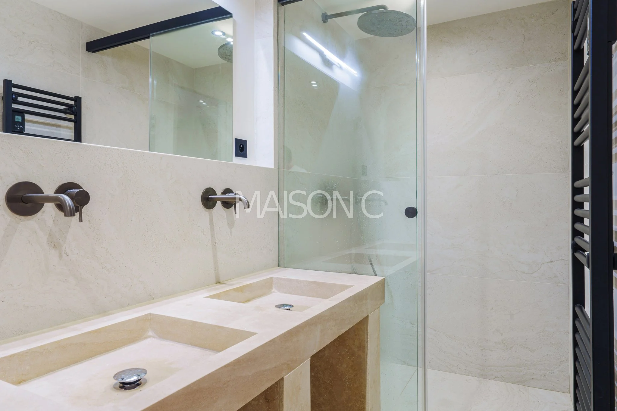 Salle de bain moderne avec double lavabo en marbre beige, miroir grand format, douche en verre avec deux pommeaux à la paroi en marbre et mur beige, radiateur noir en forme de barre, éclairage encastré au plafond.