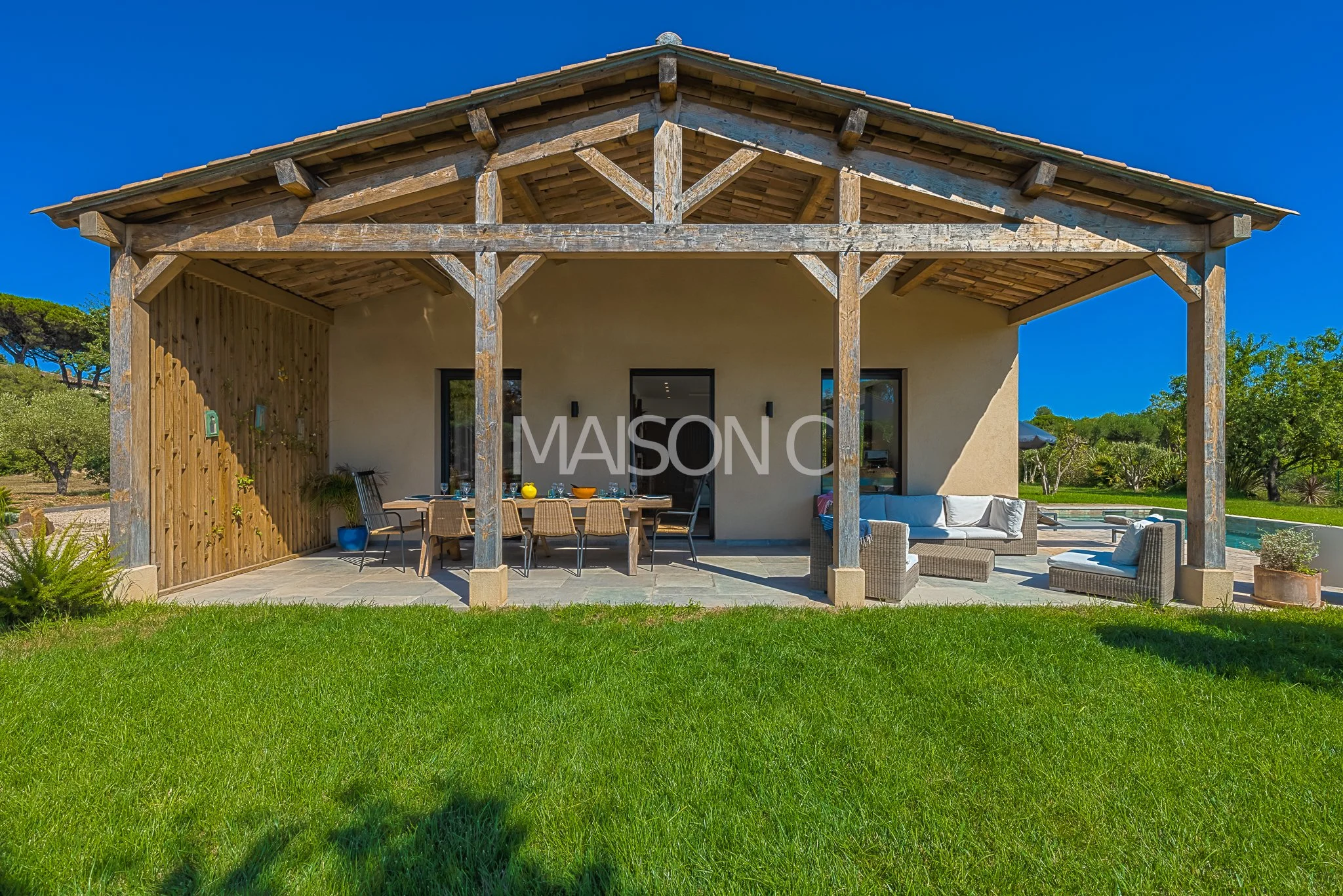 Maison de style rural avec véranda en bois, mobilier de jardin, salon et table extérieur, entourée de verdure sous un ciel bleu