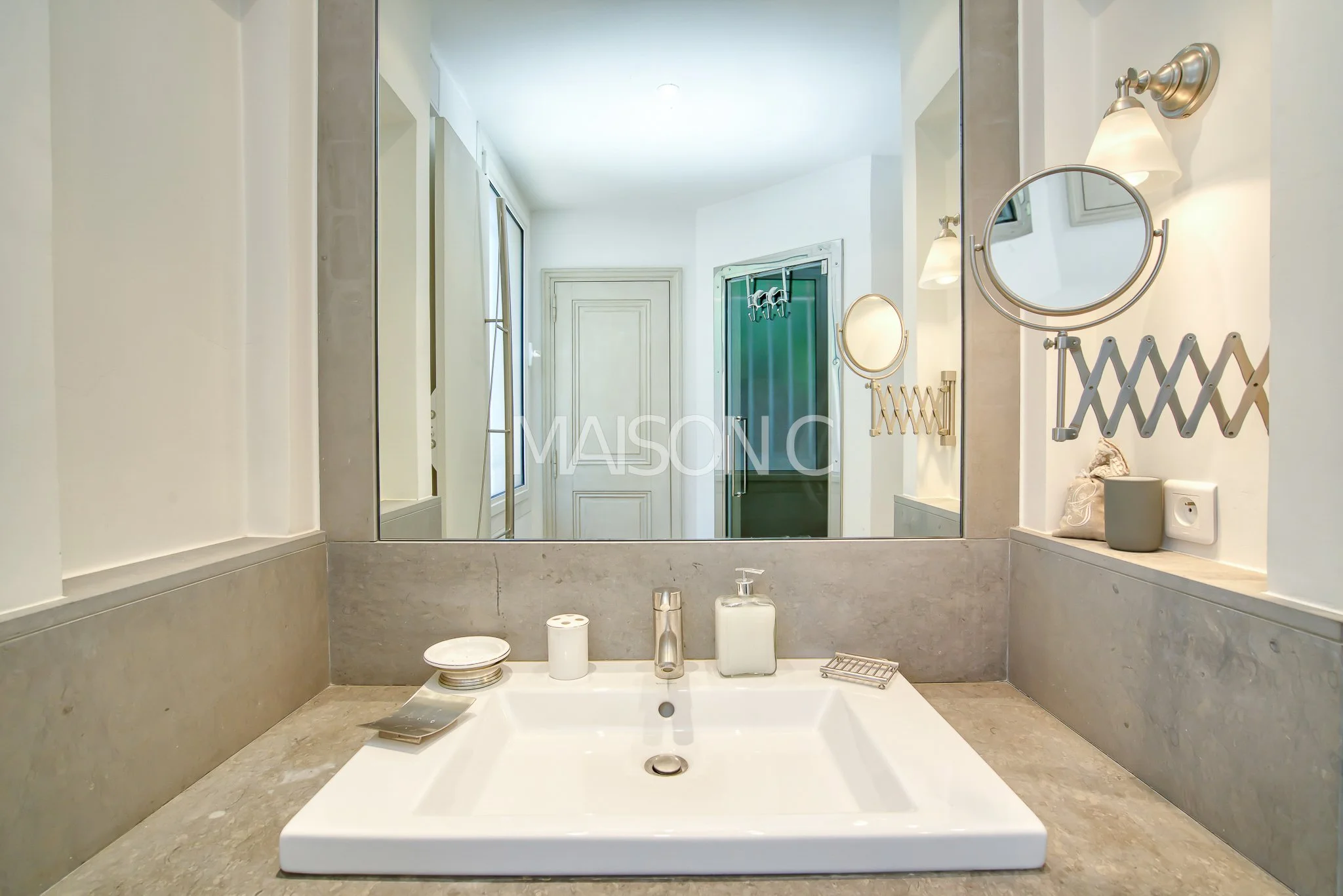 Vue intérieure d'une salle de bain moderne avec un lavabo blanc, un grand miroir, deux appliques murales, et divers accessoires de salle de bain, avec une porte et une fenêtre derrière.