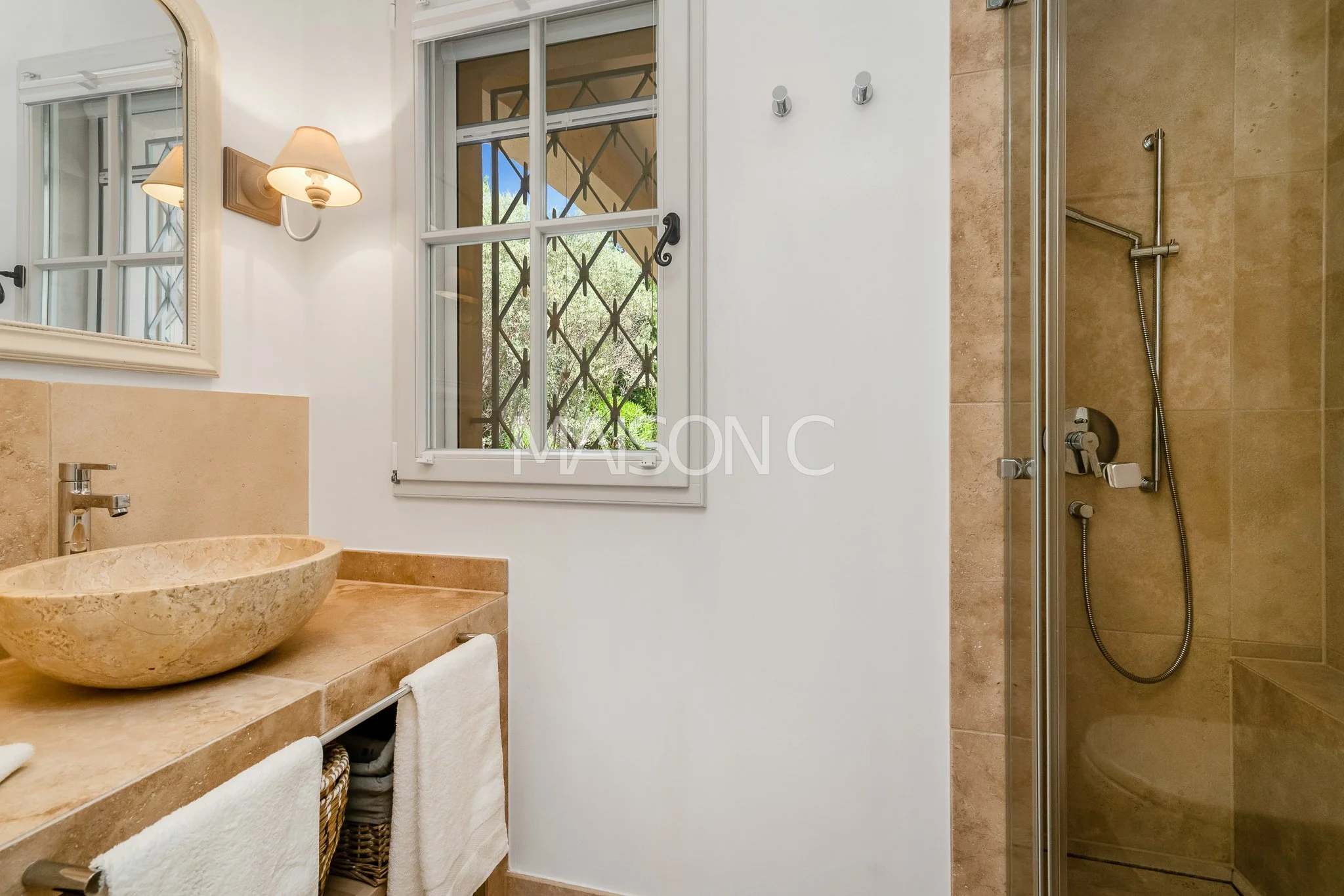 Une salle de bain avec un lavabo en pierre, un miroir, une fenêtre, et une cabine de douche avec des murs en carrelage beige.