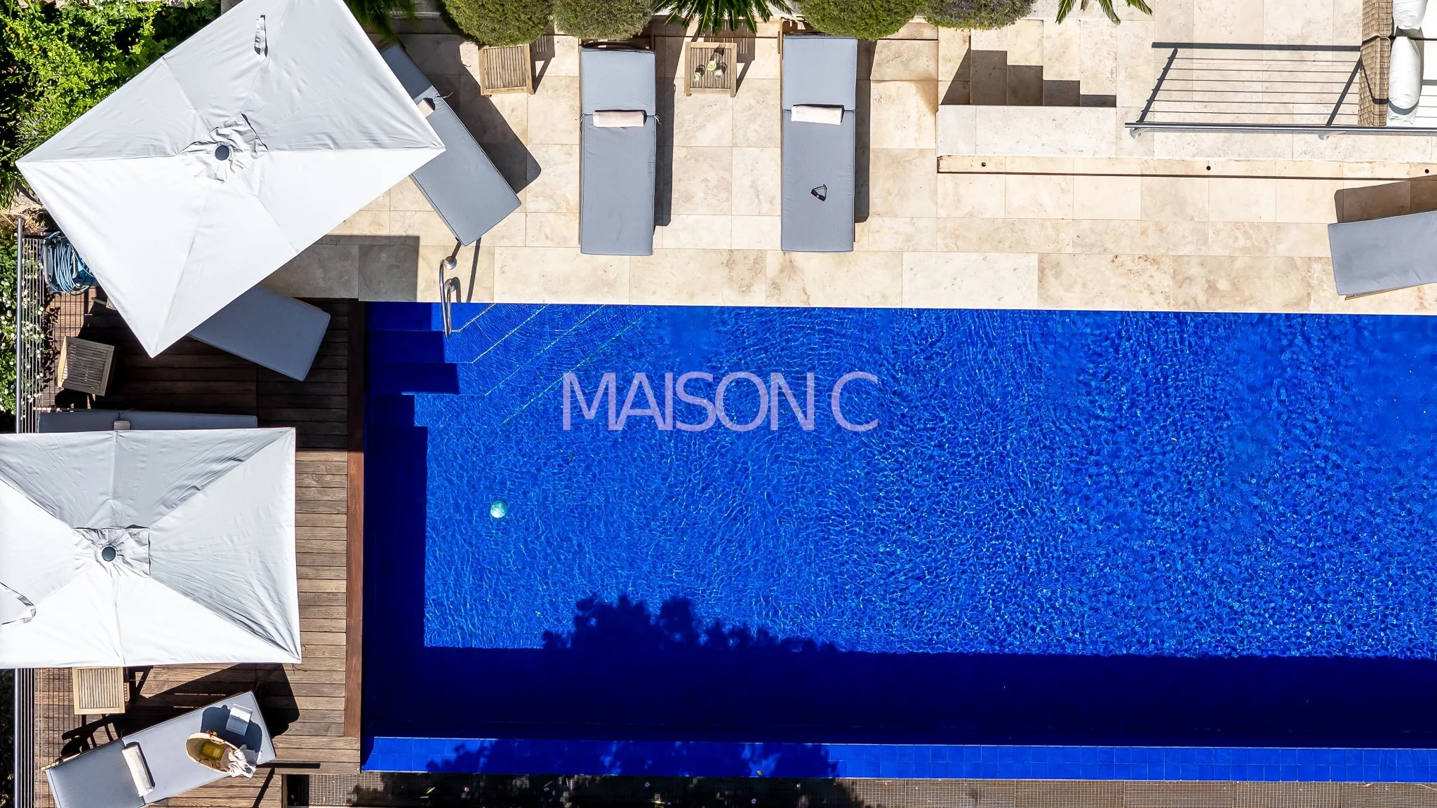 Photo aérienne d'une piscine bleue avec le texte 'MAISON C' dessus, entourée de parasols blancs, chaises longues, et une terrasse en pierre, dans un espace extérieur ensoleillé.