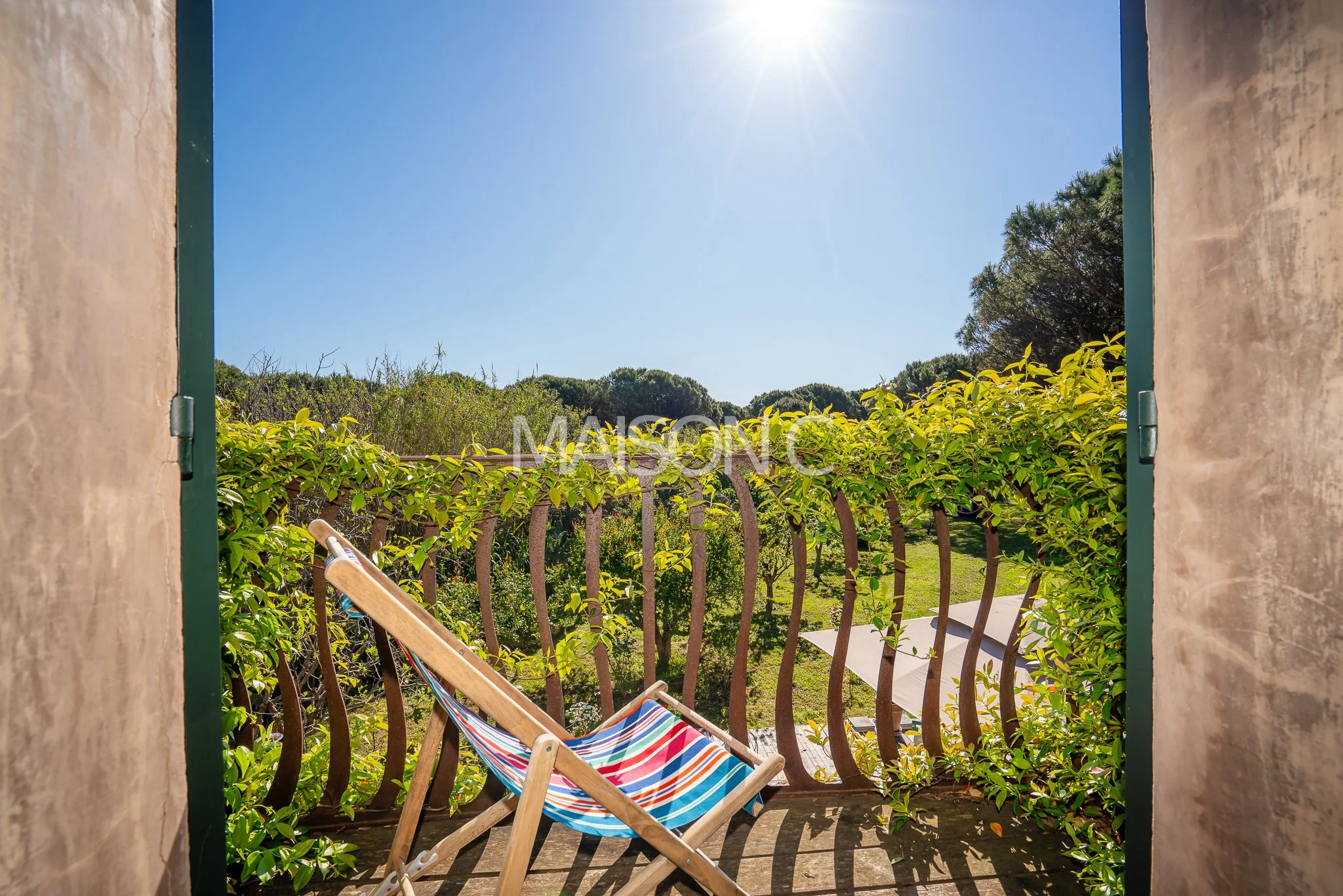 Vue depuis une fenêtre vers une terrasse et un jardin ensoleillé avec un fauteuil pliant coloré.