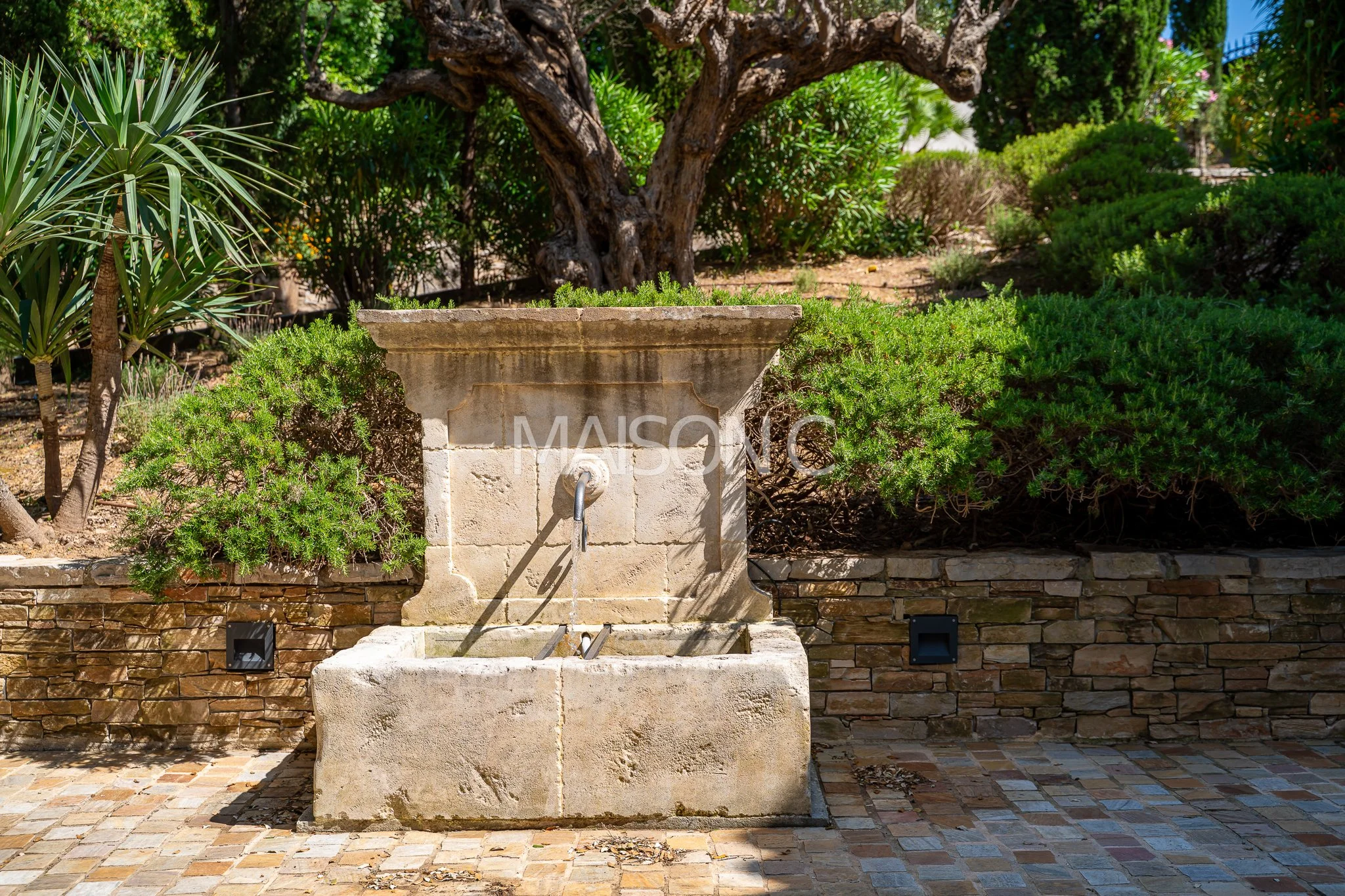 Une fontaine en pierre dans un jardin avec des buissons verts et un arbre en arrière-plan.