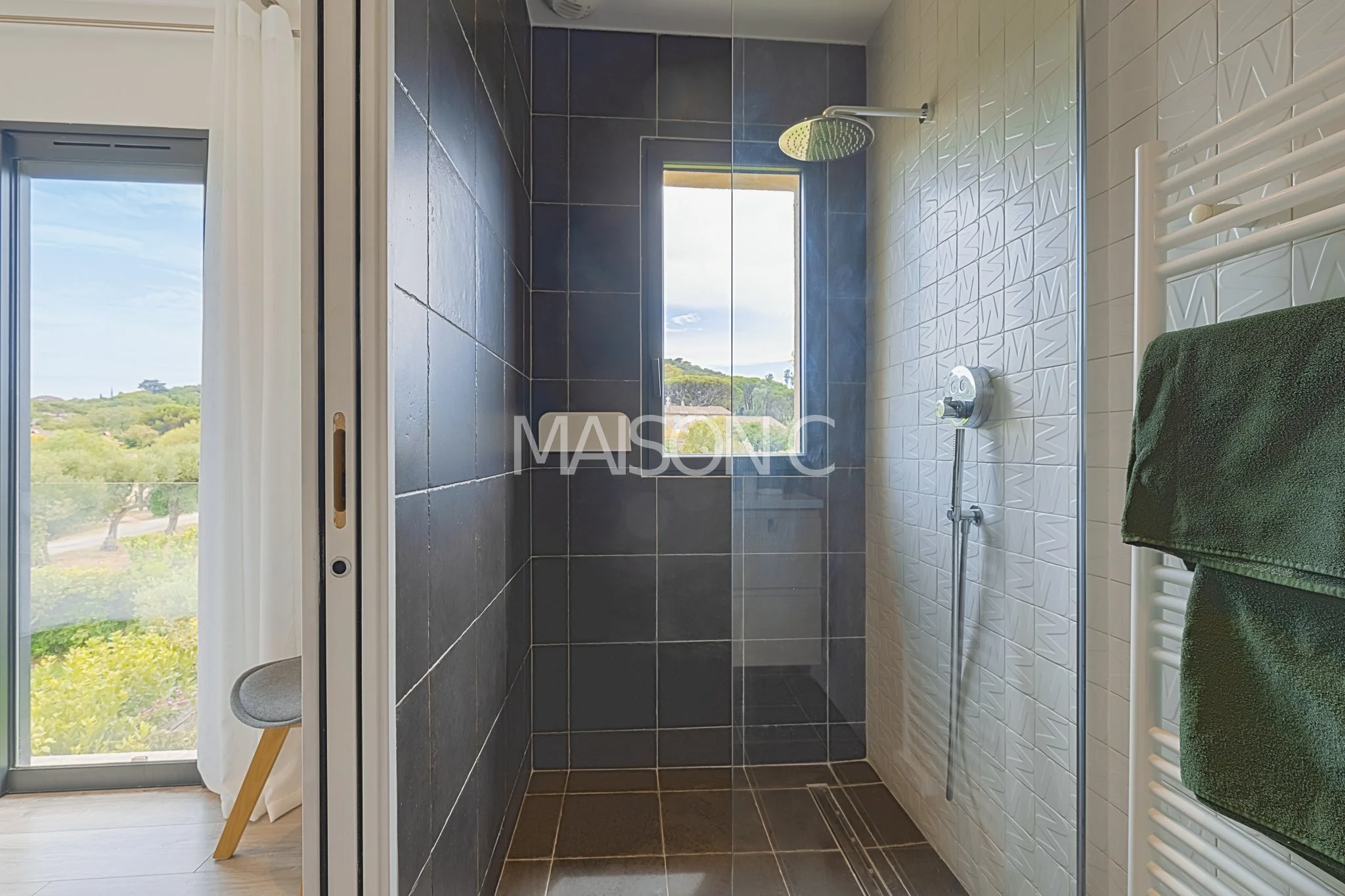 Salle de douche avec grand pommeau de douche, mur blanc avec motif en relief, fenêtre laissant entrer la lumière, serviette verte suspendue à un radiateur électrique.