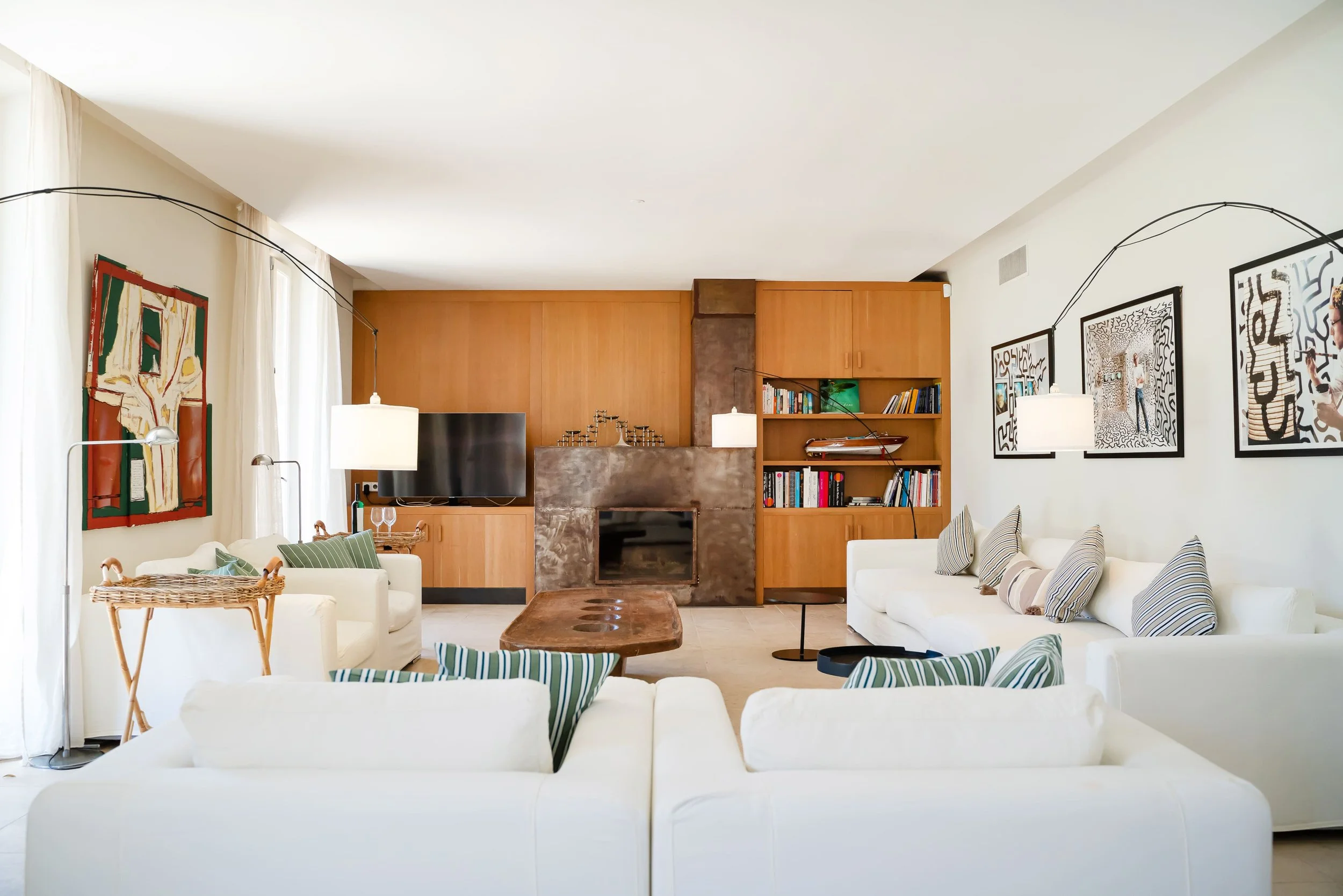 Salon moderne avec canapés blancs, tableau abstrait, cheminée, étagères en bois avec livres, télévision, lampes suspendues, et décorations artistiques.