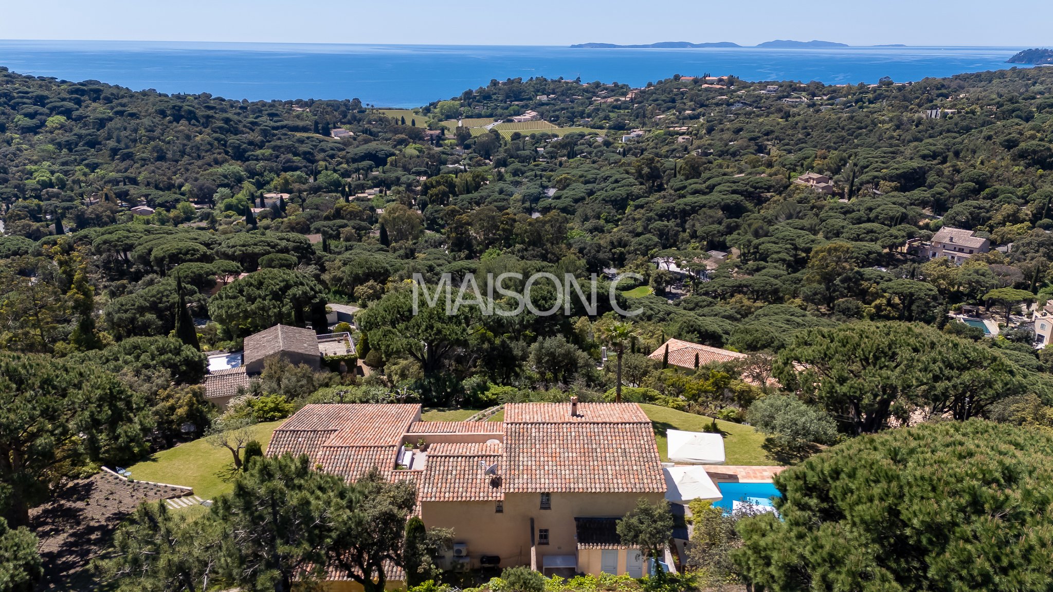 Vue aérienne d'une propriété résidentielle avec piscine au premier plan, entourée de végétation dense et de nombreuses maisons, avec la mer en arrière-plan.