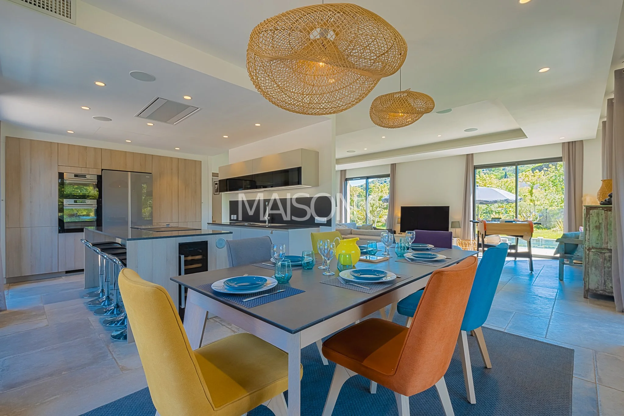Salon ouvert avec vue sur l'extérieur, mobilier coloré, table étendue avec vaisselle, cuisine moderne en arrière-plan, luminaires en rotin suspendus.