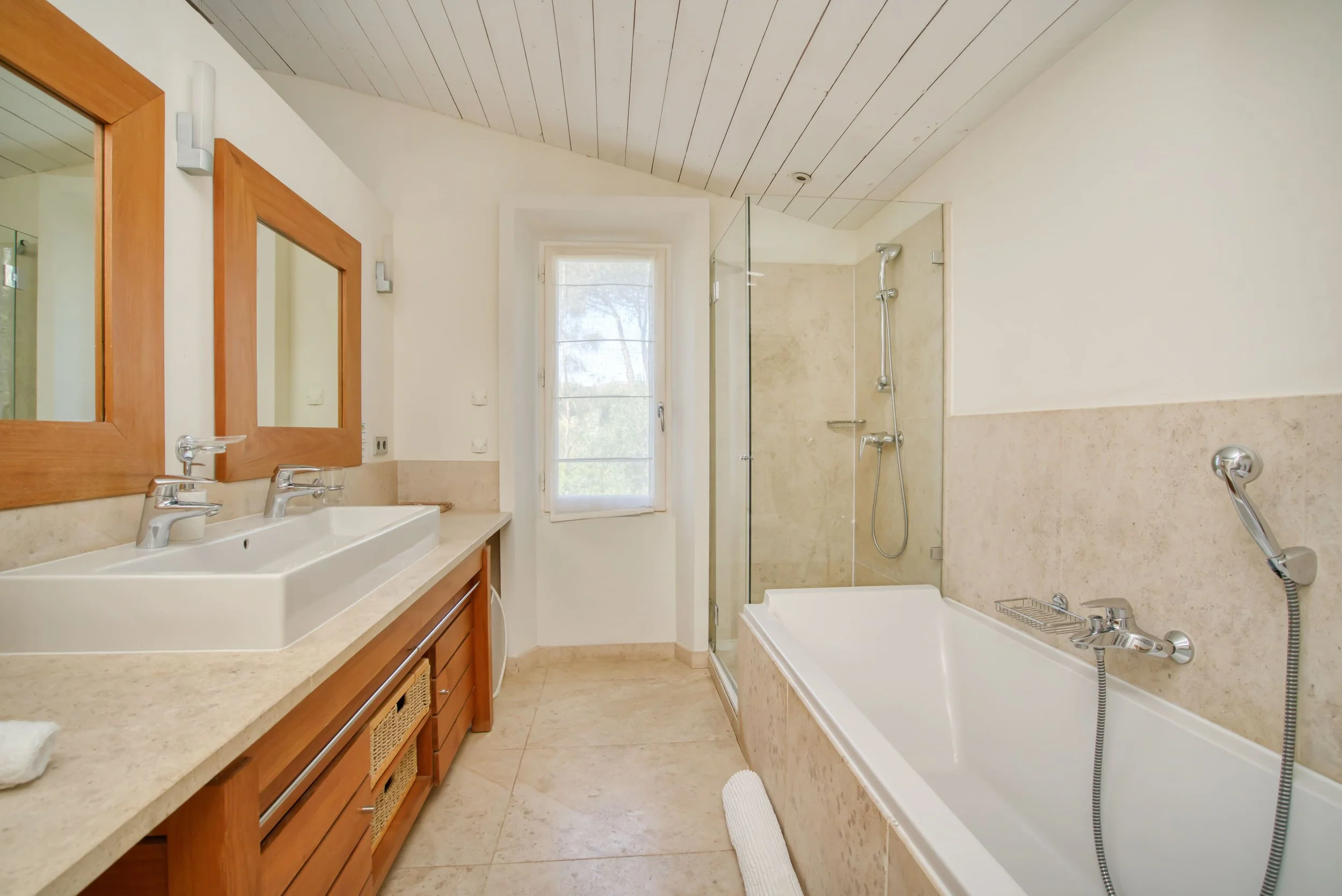 Salle de bain avec double lavabo, baignoire, douche séparée, fenêtre avec vue extérieure, murs et sol en pierre claire, meubles en bois.