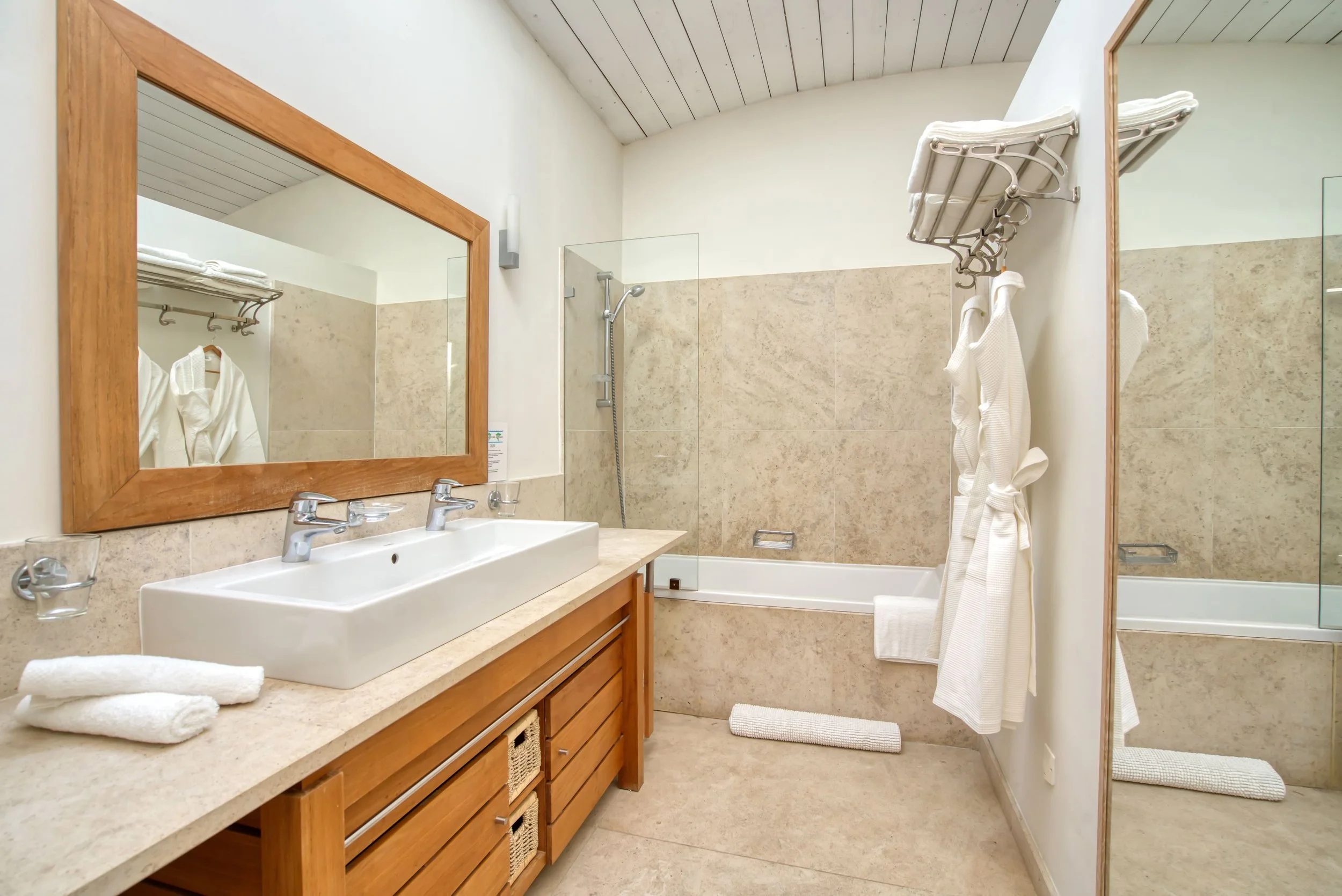 Salle de bain avec grand miroir en bois, lavabo double, douche avec paroi en verre, baignoire, serviettes blanches, tapis, et robe de chambre.