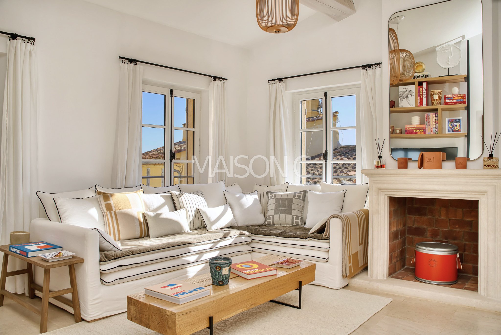 Salon lumineux avec canapé blanc, coussins variés, cheminée en pierre, grande fenêtre avec rideaux blancs, étagère avec décorations, miroir rectangulaire, table basse en bois avec livres, tapis beige, plafond avec luminaire en rotin.