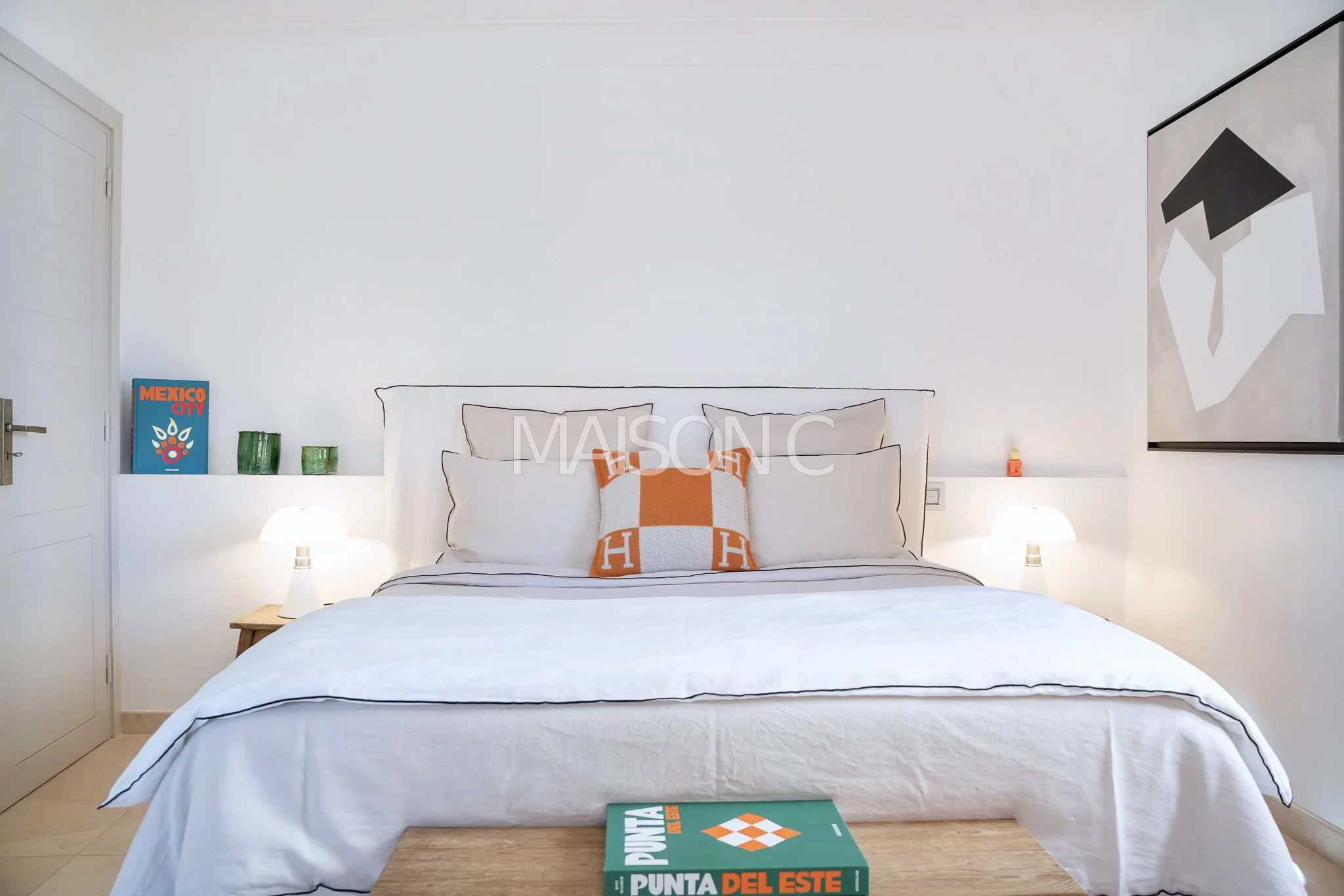 Chambre minimaliste avec un lit blanc doté de plusieurs coussins orange et blanc, deux lampes de chevet allumées, une table en bois avec un livre de jeux de société, décorations vertes et un tableau abstrait noir et blanc sur le mur.