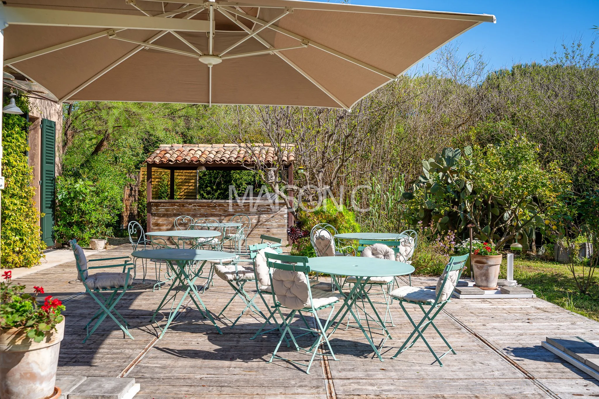 Une terrasse en bois avec plusieurs tables rondes en métal vert, des chaises avec coussins, un parasol beige, et un fond de verdure avec des plantes et un petit cabanon en bois.