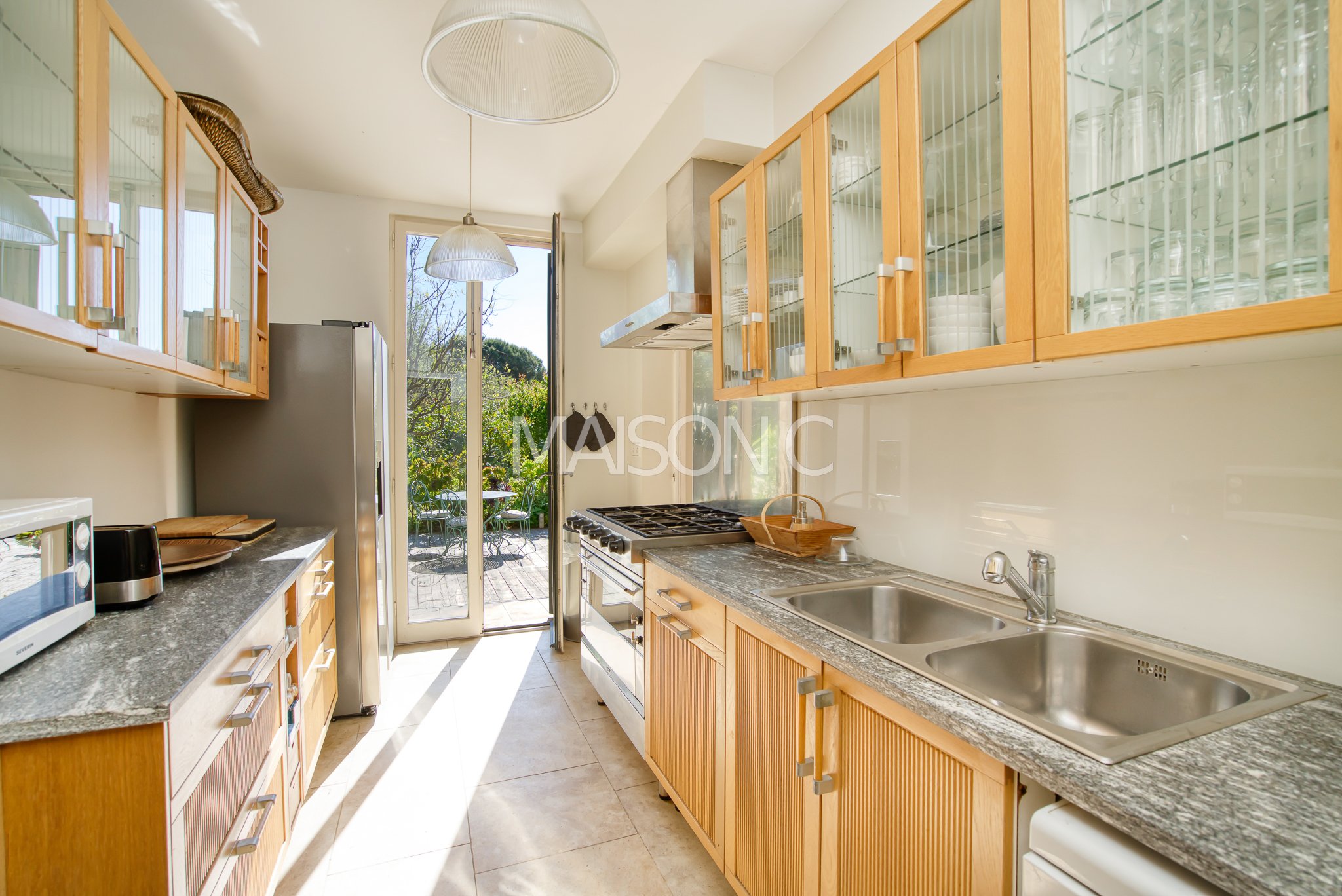 Cuisine lumineuse avec plans de travail en granit, armoires en bois, évier double, cuisinière à gaz, réfrigérateur, micro-ondes, lumière naturelle à travers une porte-fenêtre donnant sur un espace extérieur avec mobilier.