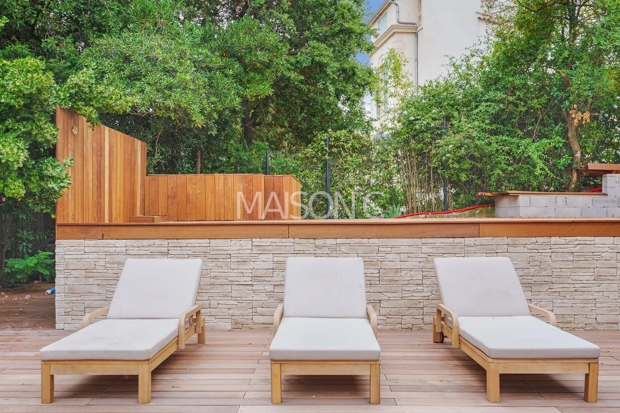 Un espace extérieur avec trois transats en bois et tissu blanc sur une terrasse en bois, avec un mur en pierre et une clôture en bois, entouré d'arbres et de plantes
