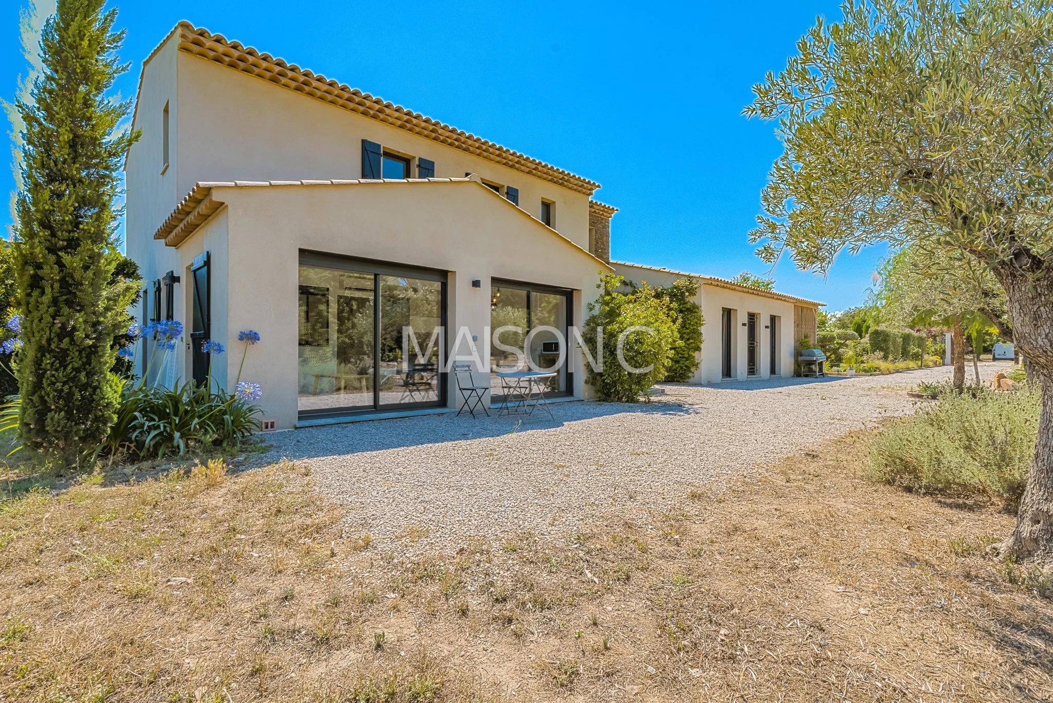 Vue extérieure d'une maison moderne avec des fenêtres en verre, un patio, des plantes et des arbres sous un ciel bleu clair.